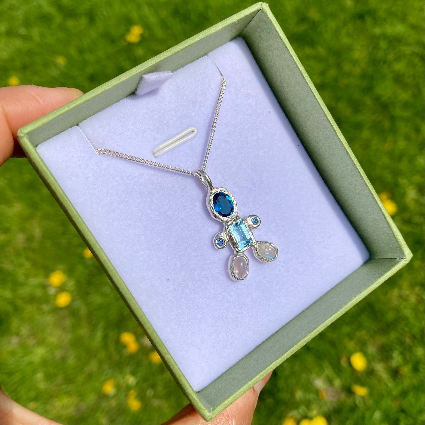 Fairy Light Pendant features light and dark blue topaz, moonstones feet. Shown in the box with green background.