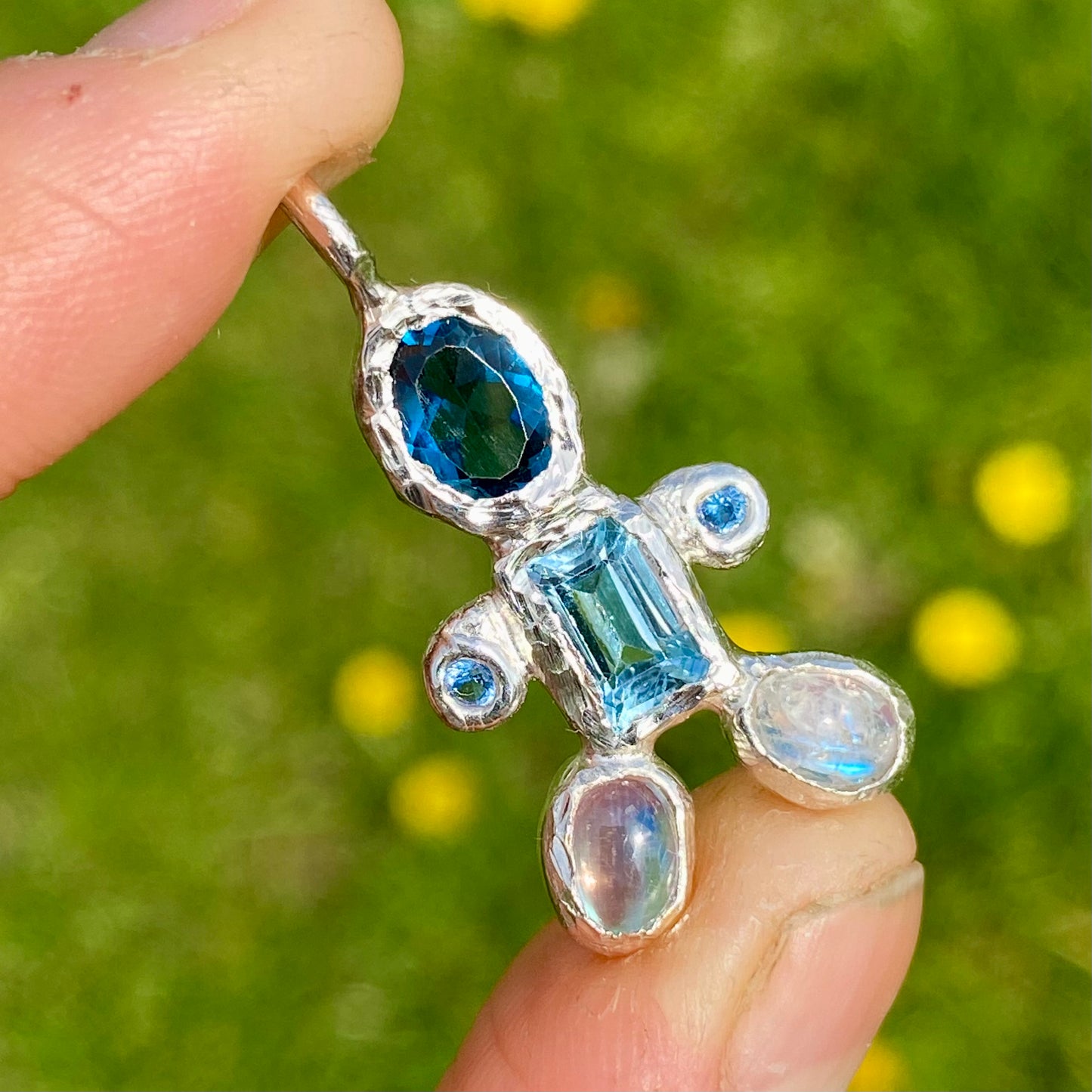 Fairy Light Pendant features light and dark blue topaz, moonstones feet. Shown in green grass as background.