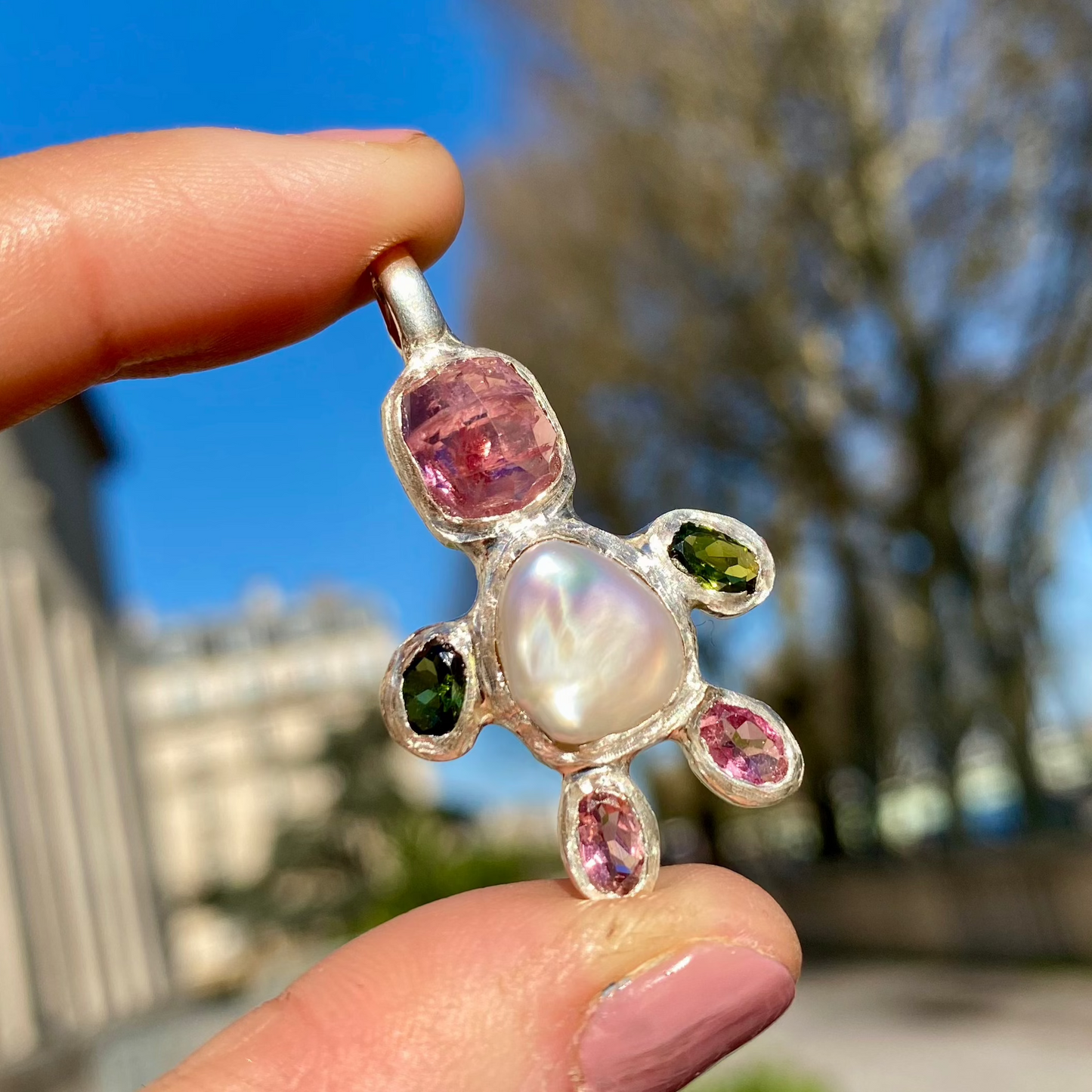 Fairy Light Pendant with white keshi pearl, pink tourmalines, & green tourmalines for the hands. Trees as background in photo