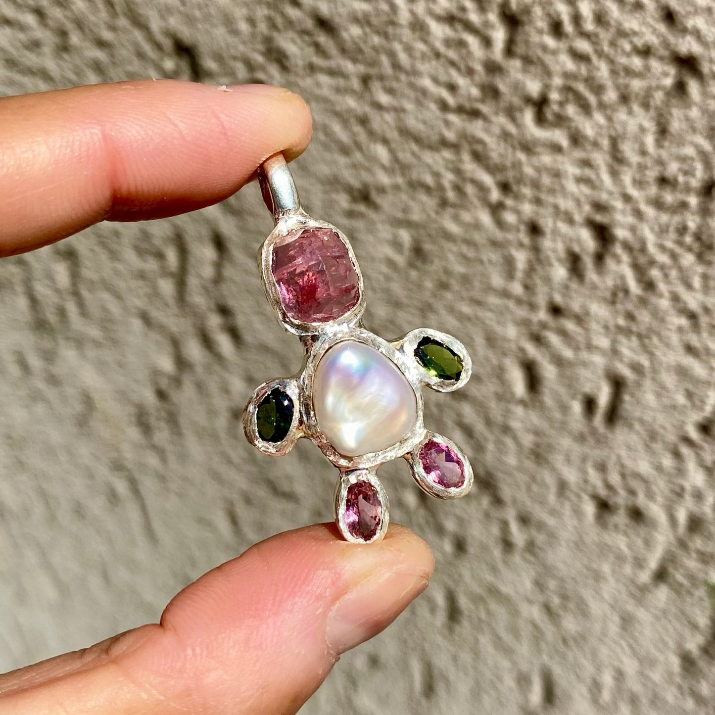 Fairy Light Pendant with white keshi pearl, pink tourmalines, & green tourmalines for the hands. Wall as background.