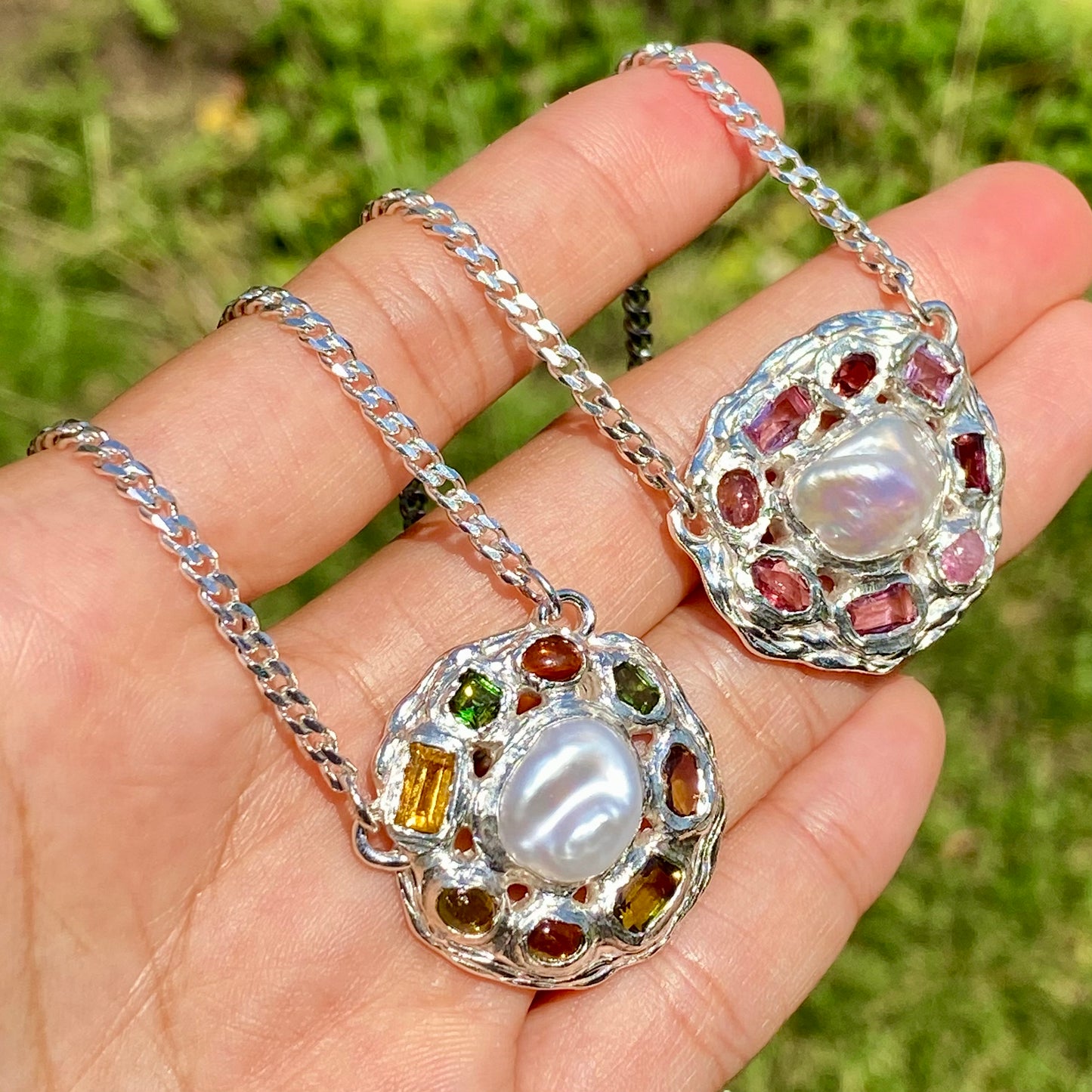 Two silver pendants with pearls and gemstones held in a hand against a grassy background