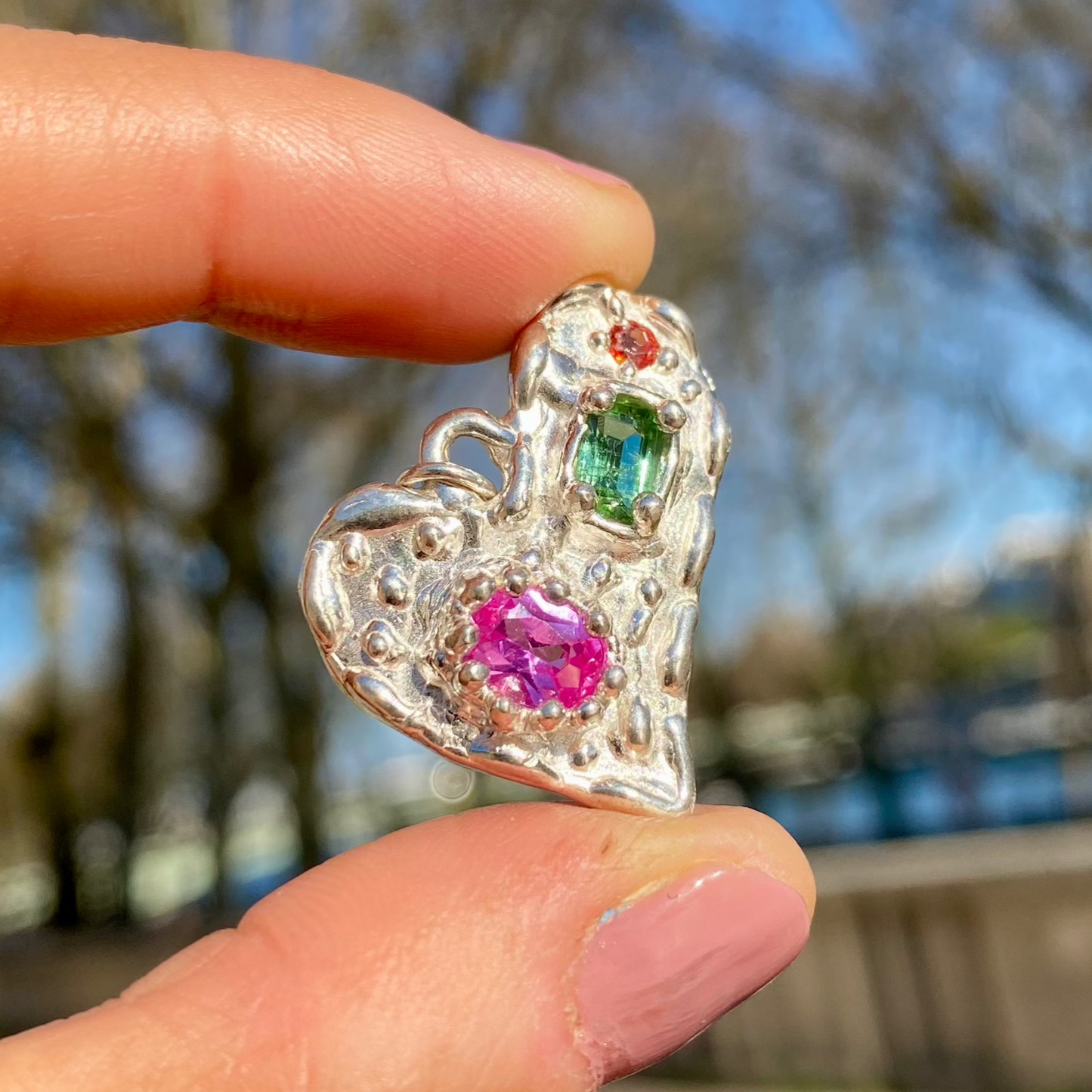 Handmade by Lia from Lia Makes Jewellery, this sterling silver heart-shaped pendant features a unique combination of colourful stones like tourmalines, sapphires, citrine, pink and blue topaz, and tiny synthetic gems, shown in a close-up to highlight the vibrant setting.