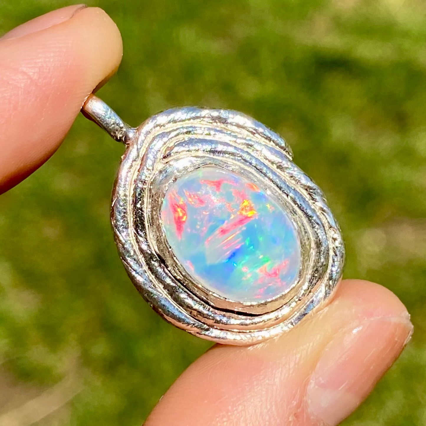 Opal pendant with silver setting held between fingers against a blurred green background
