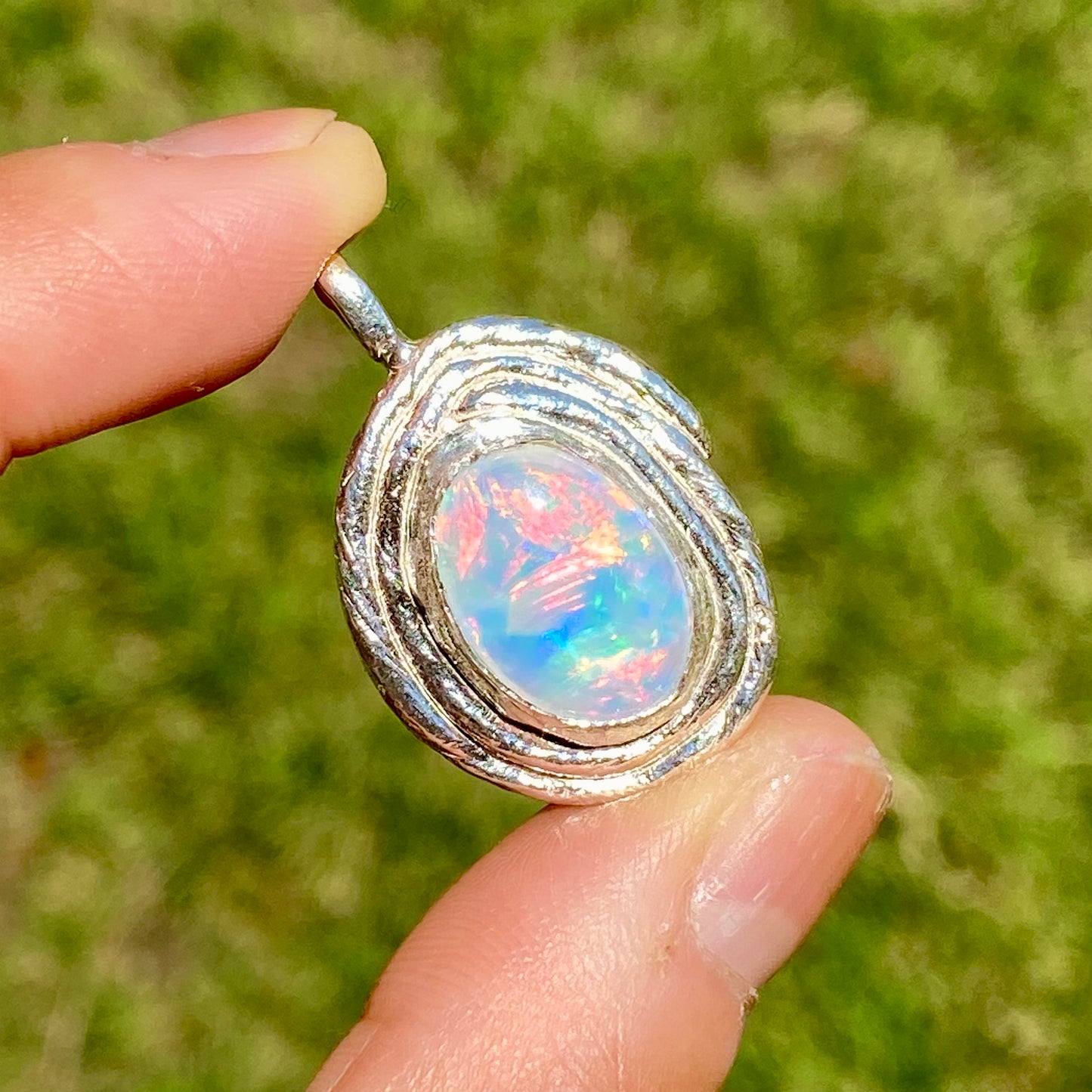 Opal pendant with silver setting held between fingers against a blurred green background