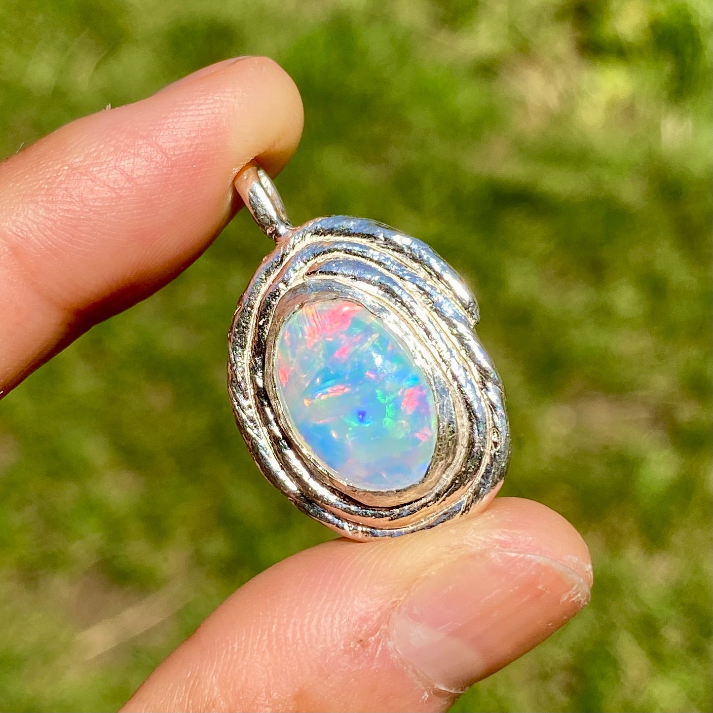 Opal pendant in silver setting held between fingers with a blurred green background