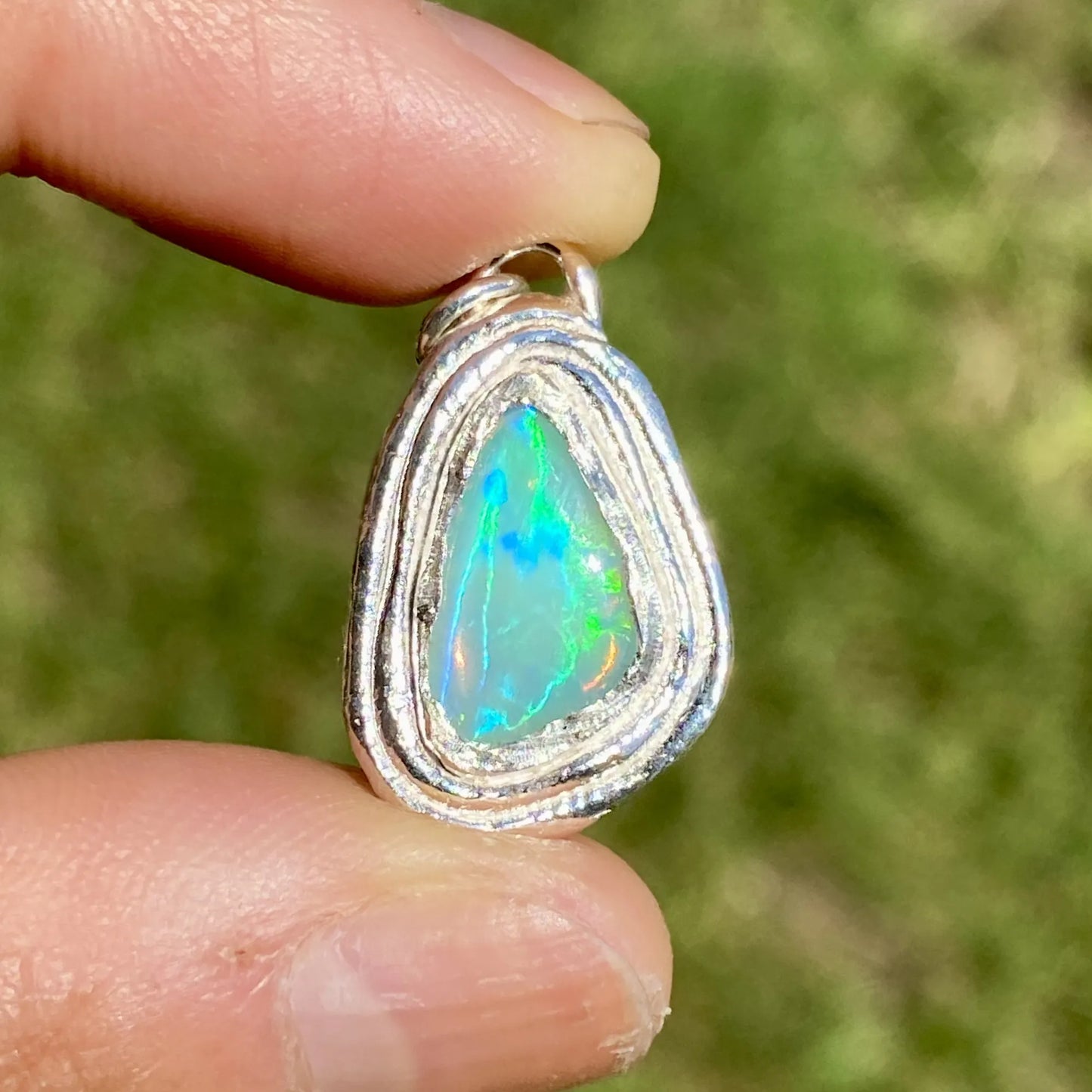 Opal pendant with silver setting held between fingers against a blurred green background