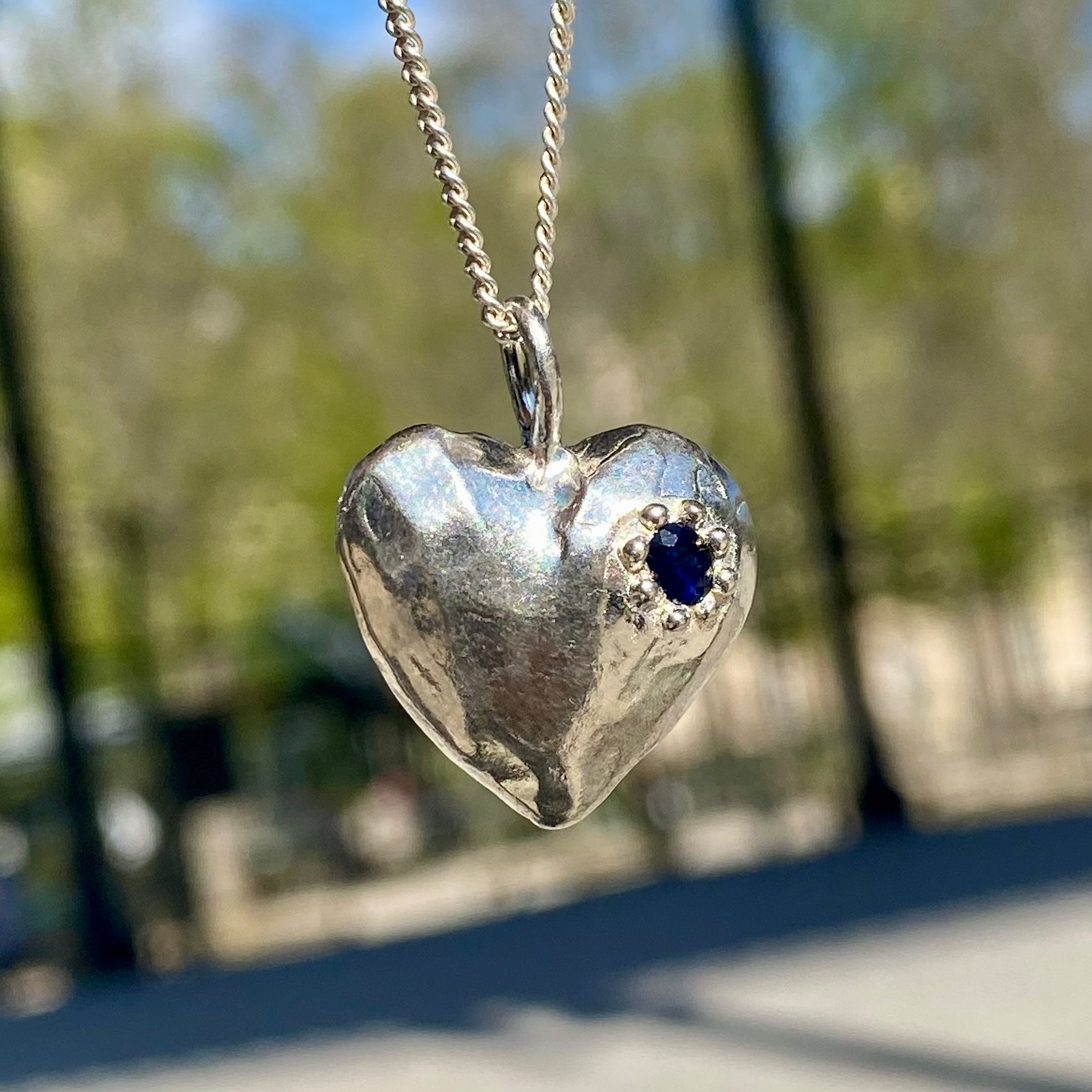 The Petite Heart pendant is a heart-shaped piece, handmade by me in sterling silver. Each pendant features a sapphire stone and is available in four colours: yellow, pink, orange, or blue.