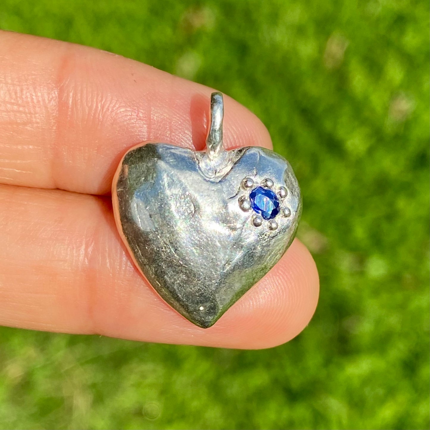 The Petite Heart pendant is a heart-shaped piece, handmade by me in sterling silver. Each pendant features a sapphire stone and is available in four colours: yellow, pink, orange, or blue.