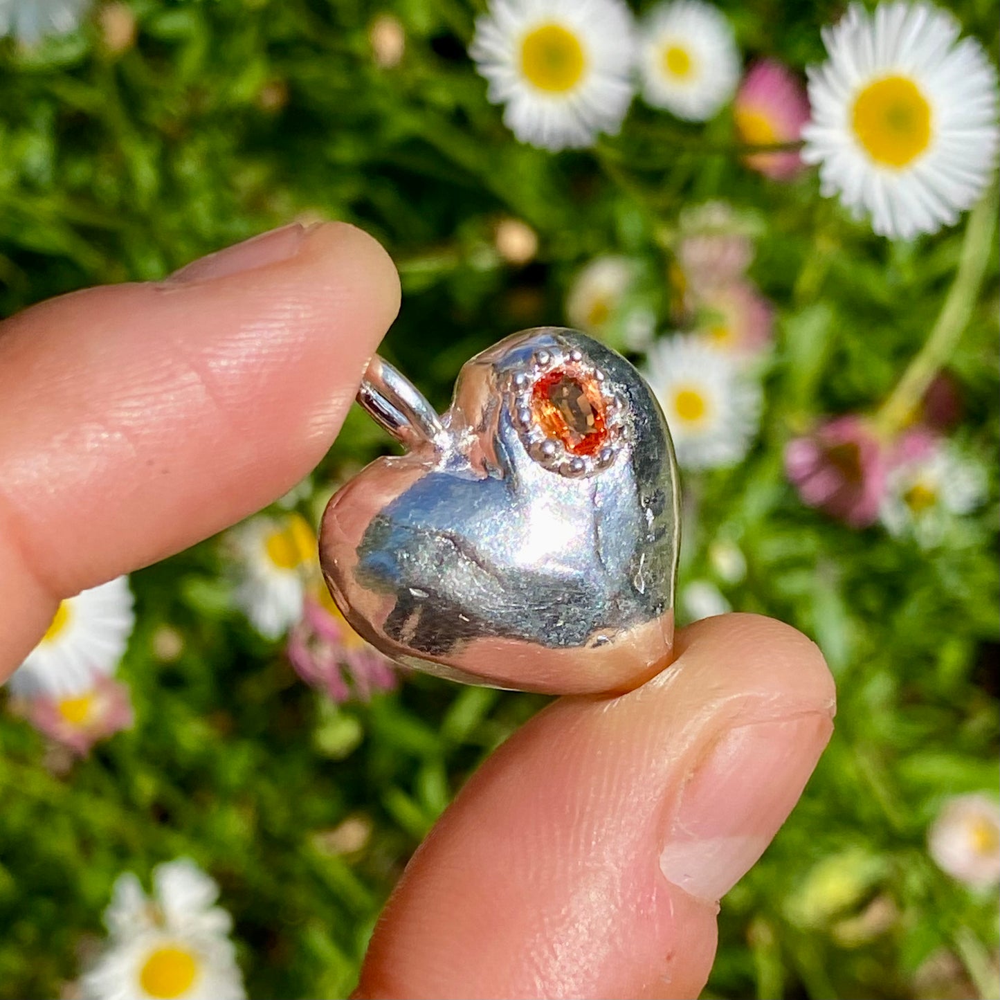 The Petite Heart pendant is a heart-shaped piece, handmade by me in sterling silver. Each pendant features a sapphire stone and is available in four colours: yellow, pink, orange, or blue.