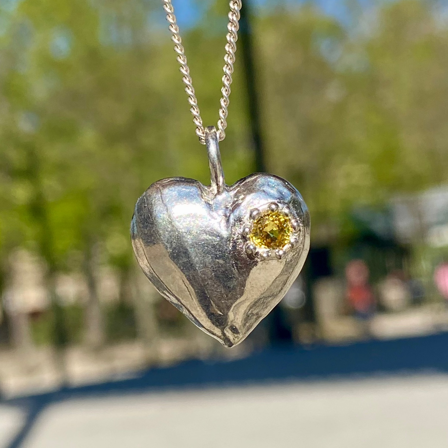 The Petite Heart pendant is a heart-shaped piece, handmade by me in sterling silver. Each pendant features a sapphire stone and is available in four colours: yellow, pink, orange, or blue.