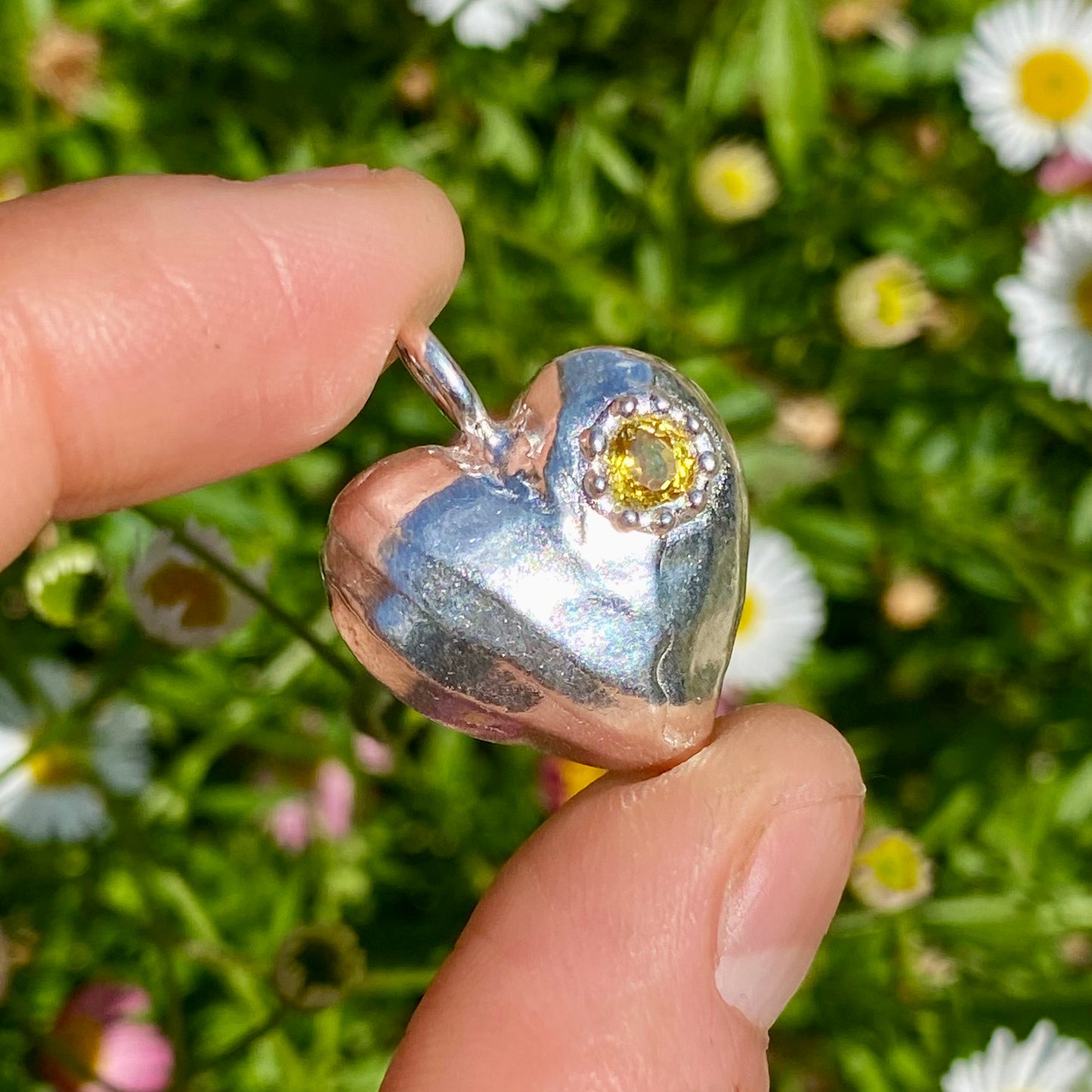 The Petite Heart pendant is a heart-shaped piece, handmade by me in sterling silver. Each pendant features a sapphire stone and is available in four colours: yellow, pink, orange, or blue.