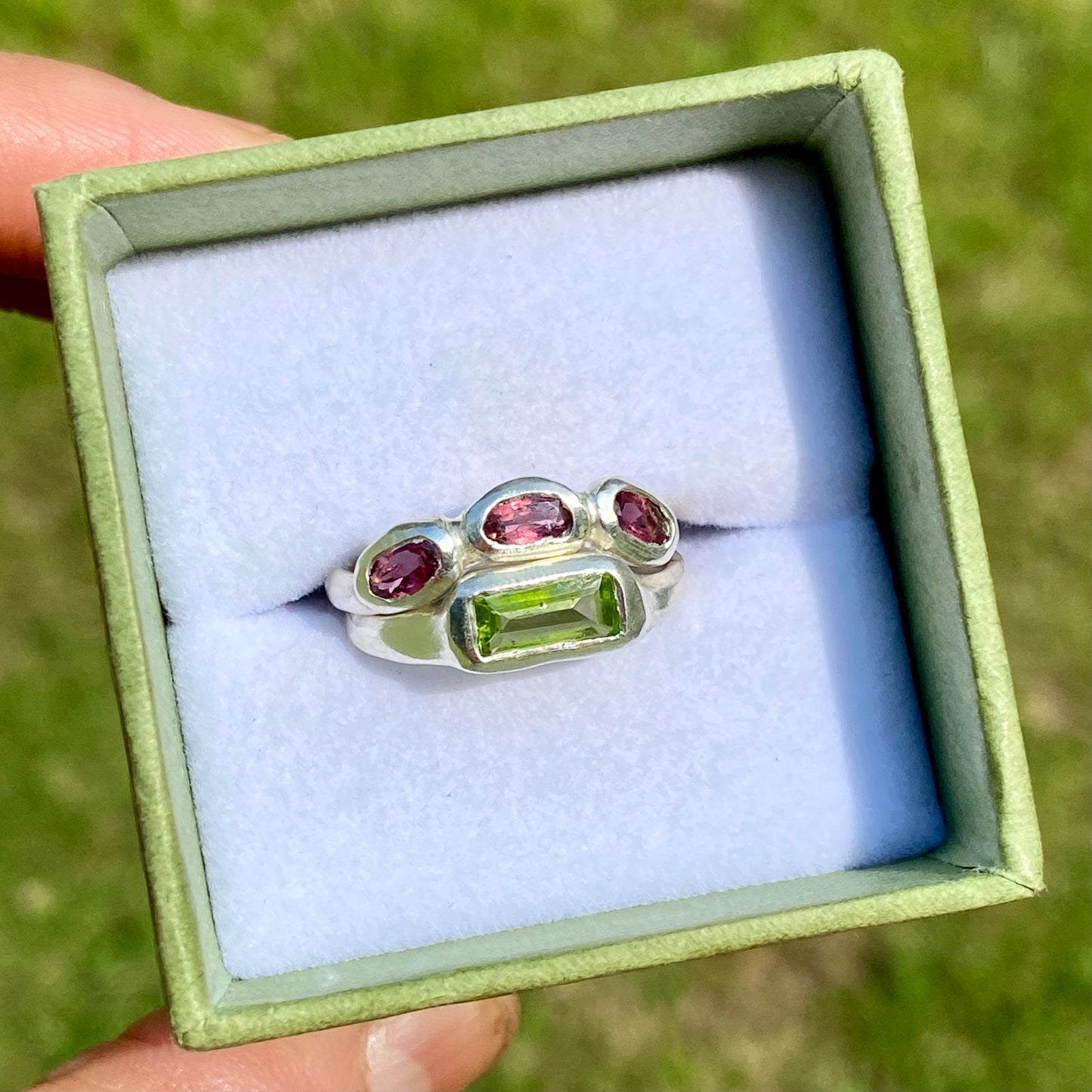 The Spring Blossom ring stack comes as a set of two. One ring features three pink tourmaline stones, and the other has a long green rectangular peridot stone. Handmade from sterling silver, these stacking rings are available in one size: O ½ (US 7 ¼).