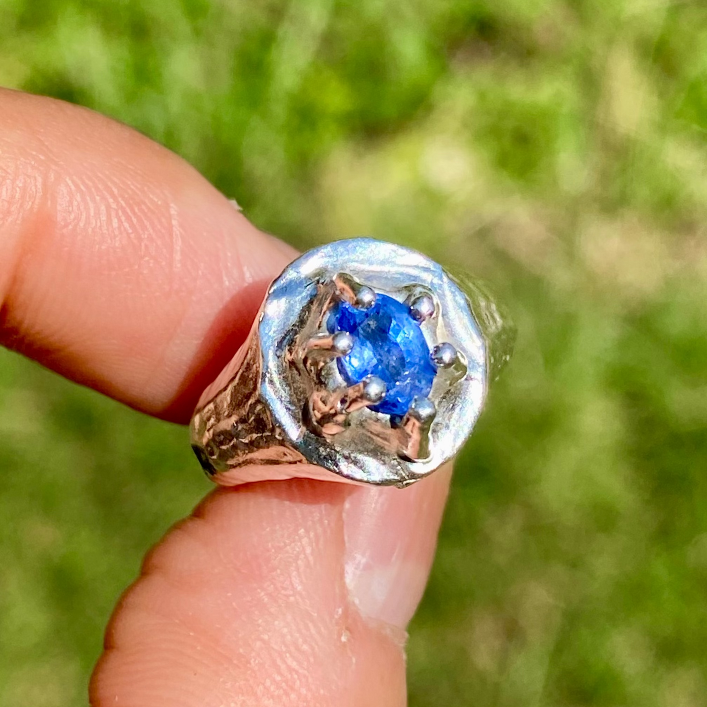 Silver ring with a blue gemstone held on a finger against a blurred green background