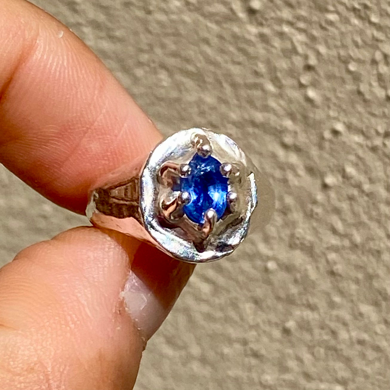 Silver ring with a blue gemstone held between two fingers against a neutral background
