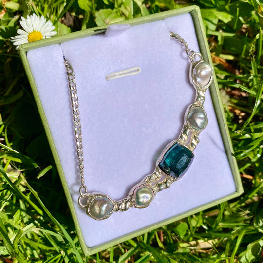 This handcrafted Aglaonema Bar pendant features a bar design inspired by the Aglaonema plant, also known as the Chinese Evergreen. The pendant comes with a big green tourmaline gemstone in the center, surrounded by four grey keshi pearls.