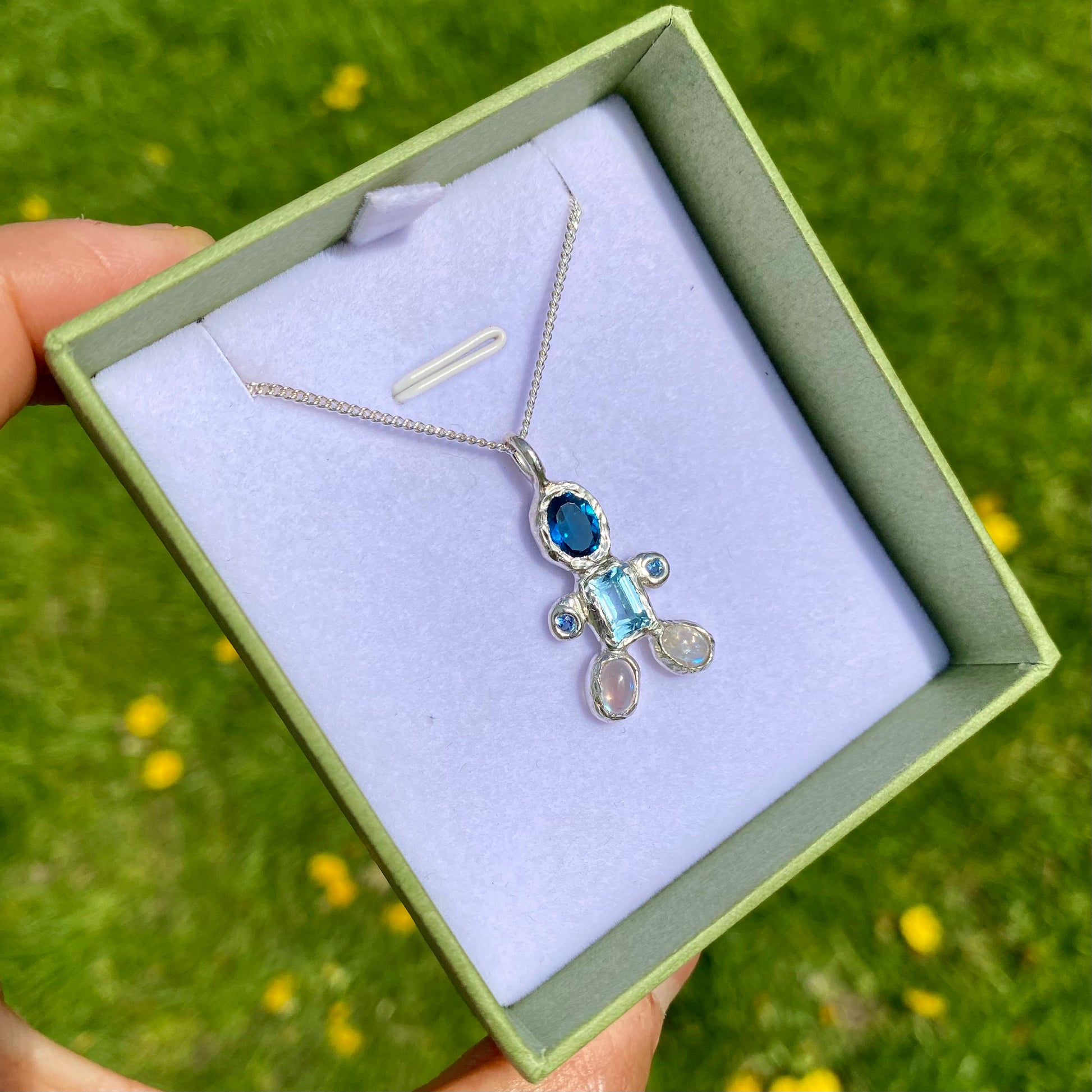 Fairy Light Pendant features light and dark blue topaz, moonstones feet. Shown in the box with green background.