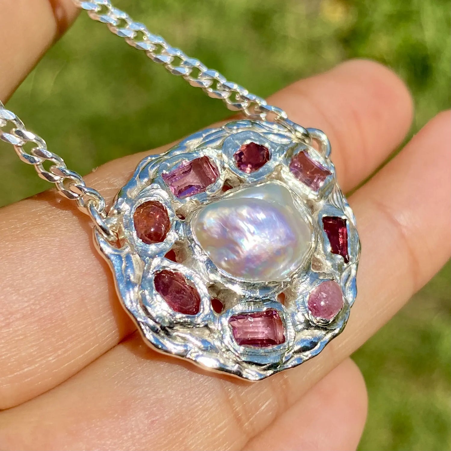 Silver pendant with a central pearl and red gemstones held in a hand against a blurred green background