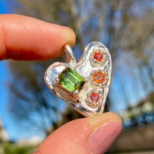 Handmade by Lia from Lia Makes Jewellery, this sterling silver heart-shaped pendant features a unique combination of colourful stones like tourmalines, sapphires, citrine, pink and blue topaz, and tiny synthetic gems, shown in a close-up to highlight the vibrant setting.
