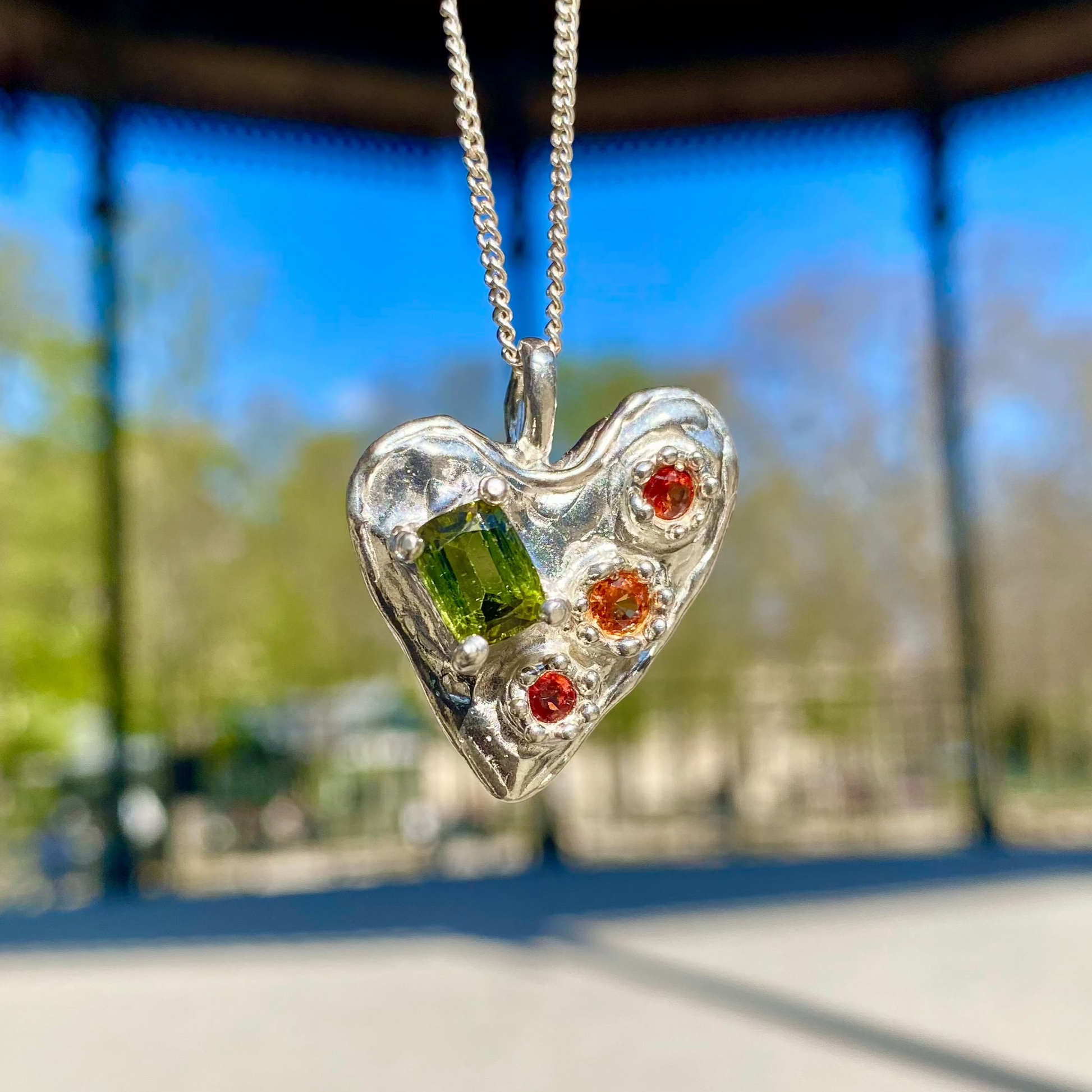 Handmade by Lia from Lia Makes Jewellery, this sterling silver heart-shaped pendant features a unique combination of colourful stones like tourmalines, sapphires, citrine, pink and blue topaz, and tiny synthetic gems, shown in a close-up to highlight the vibrant setting.
