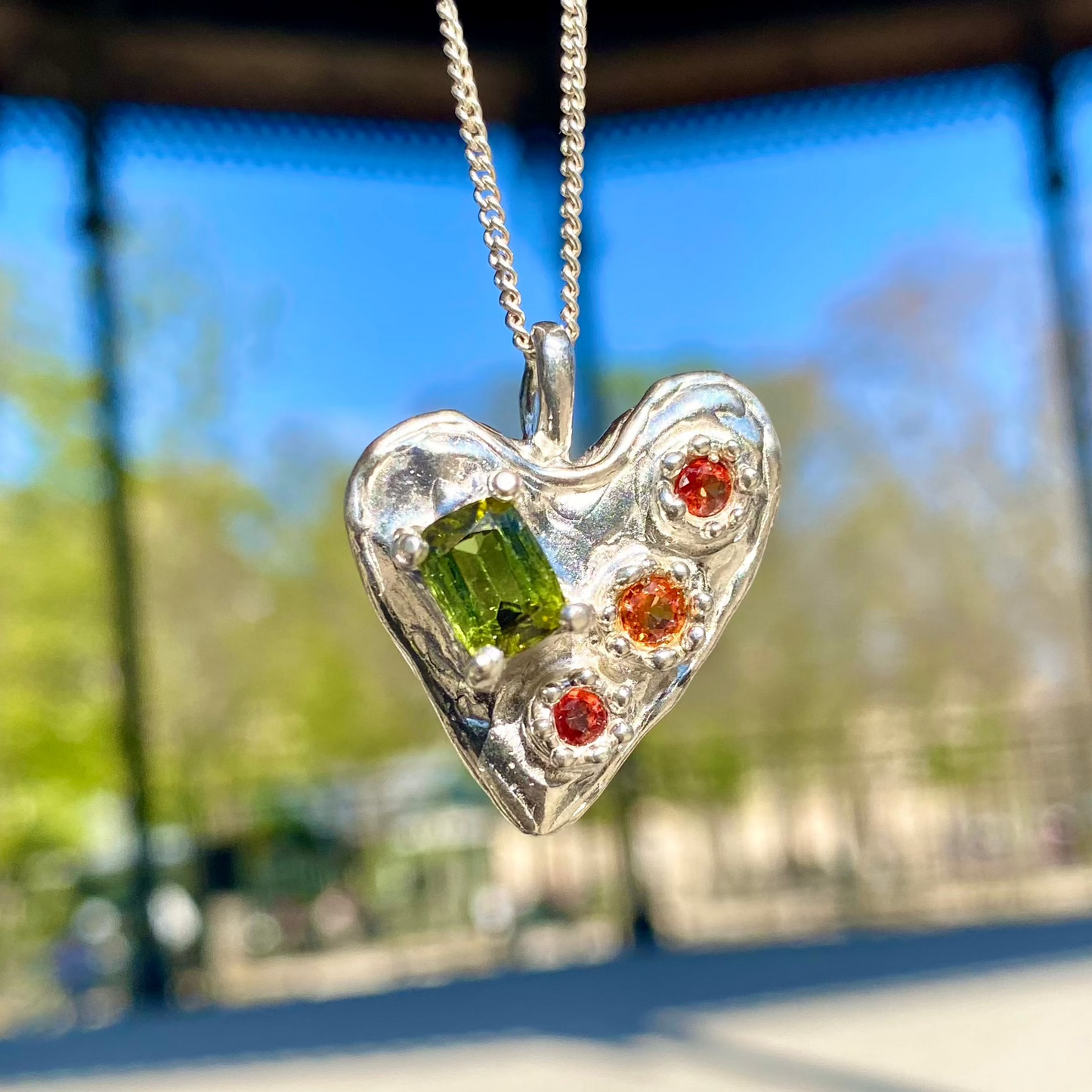Handmade by Lia from Lia Makes Jewellery, this sterling silver heart-shaped pendant features a unique combination of colourful stones like tourmalines, sapphires, citrine, pink and blue topaz, and tiny synthetic gems, shown in a close-up to highlight the vibrant setting.
