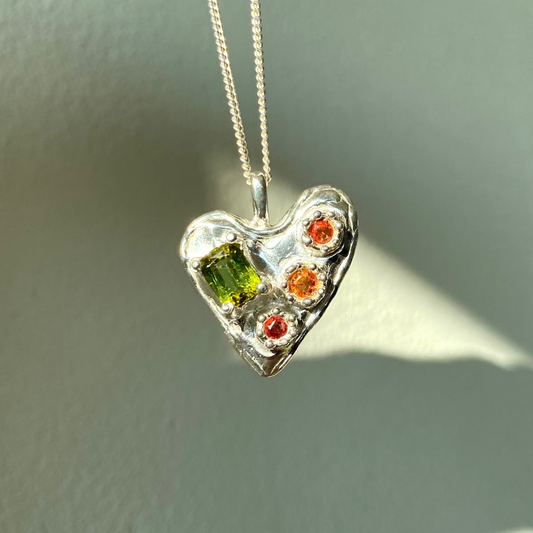 Handmade by Lia from Lia Makes Jewellery, this sterling silver heart-shaped pendant features a unique combination of colourful stones like tourmalines, sapphires, citrine, pink and blue topaz, and tiny synthetic gems, shown in a close-up to highlight the vibrant setting.

