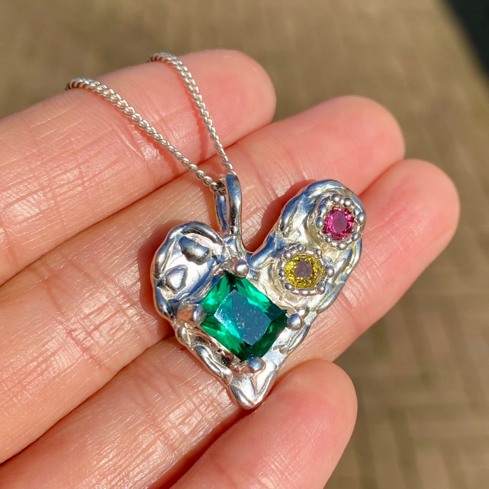 Handmade by Lia from Lia Makes Jewellery, this sterling silver heart-shaped pendant features a unique combination of colourful stones like tourmalines, sapphires, citrine, pink and blue topaz, and tiny synthetic gems, shown in a close-up to highlight the vibrant setting.
