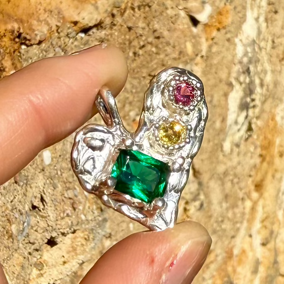 Handmade by Lia from Lia Makes Jewellery, this sterling silver heart-shaped pendant features a unique combination of colourful stones like tourmalines, sapphires, citrine, pink and blue topaz, and tiny synthetic gems, shown in a close-up to highlight the vibrant setting.
