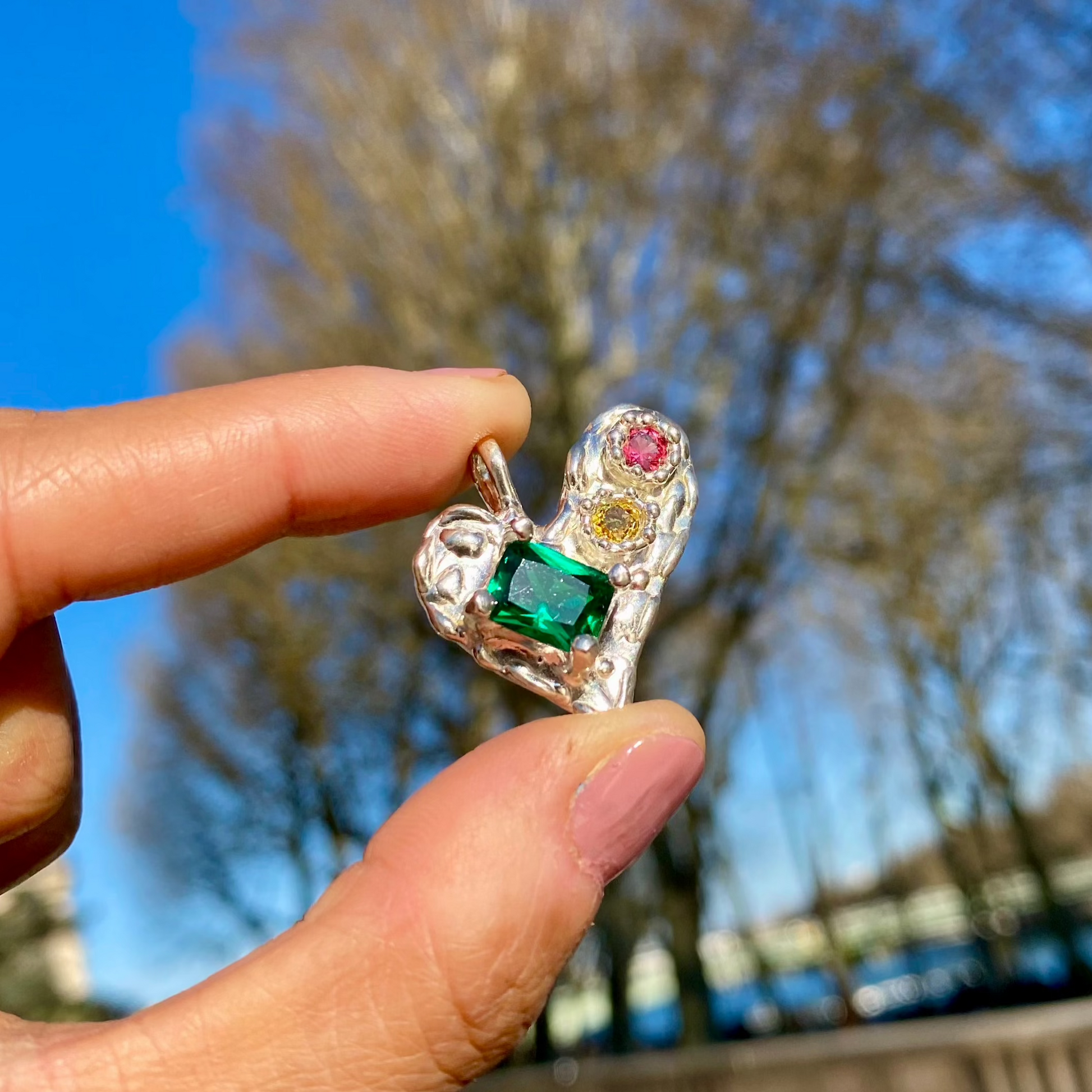 Handmade by Lia from Lia Makes Jewellery, this sterling silver heart-shaped pendant features a unique combination of colourful stones like tourmalines, sapphires, citrine, pink and blue topaz, and tiny synthetic gems, shown in a close-up to highlight the vibrant setting.
