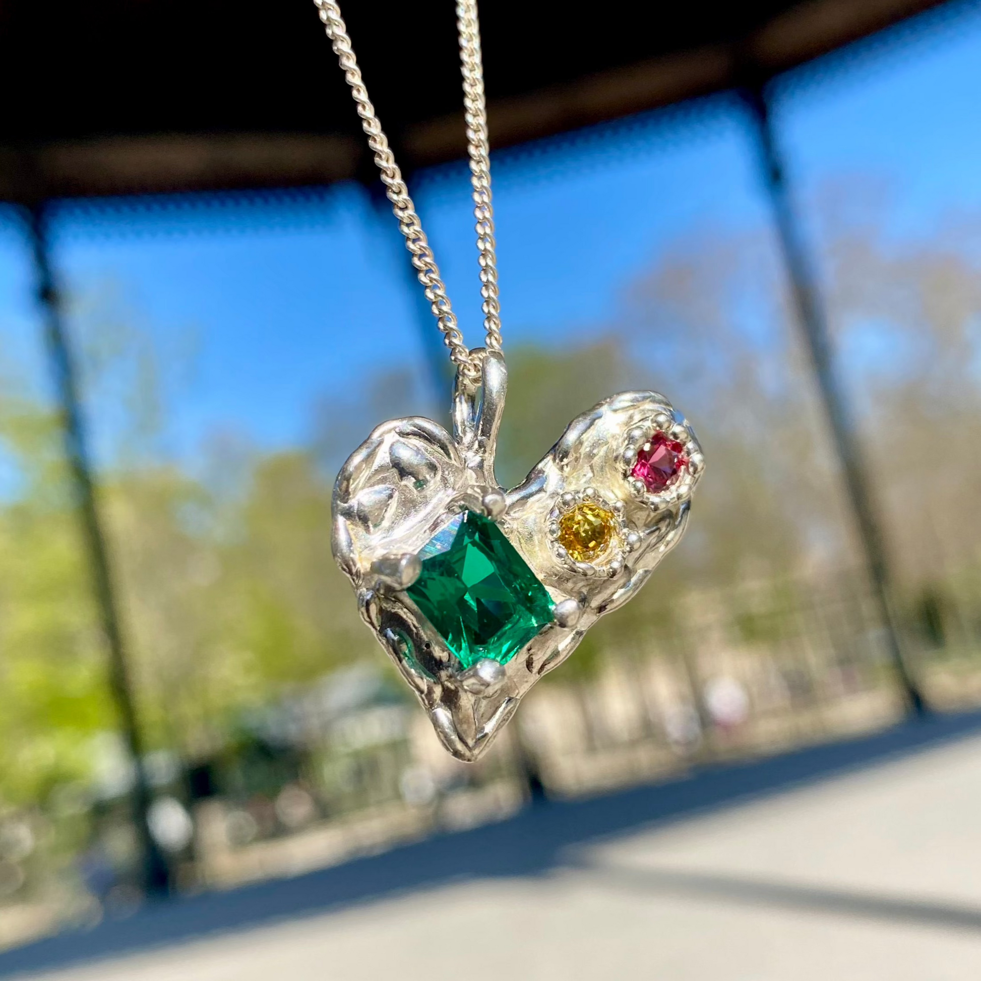 Handmade by Lia from Lia Makes Jewellery, this sterling silver heart-shaped pendant features a unique combination of colourful stones like tourmalines, sapphires, citrine, pink and blue topaz, and tiny synthetic gems, shown in a close-up to highlight the vibrant setting.
