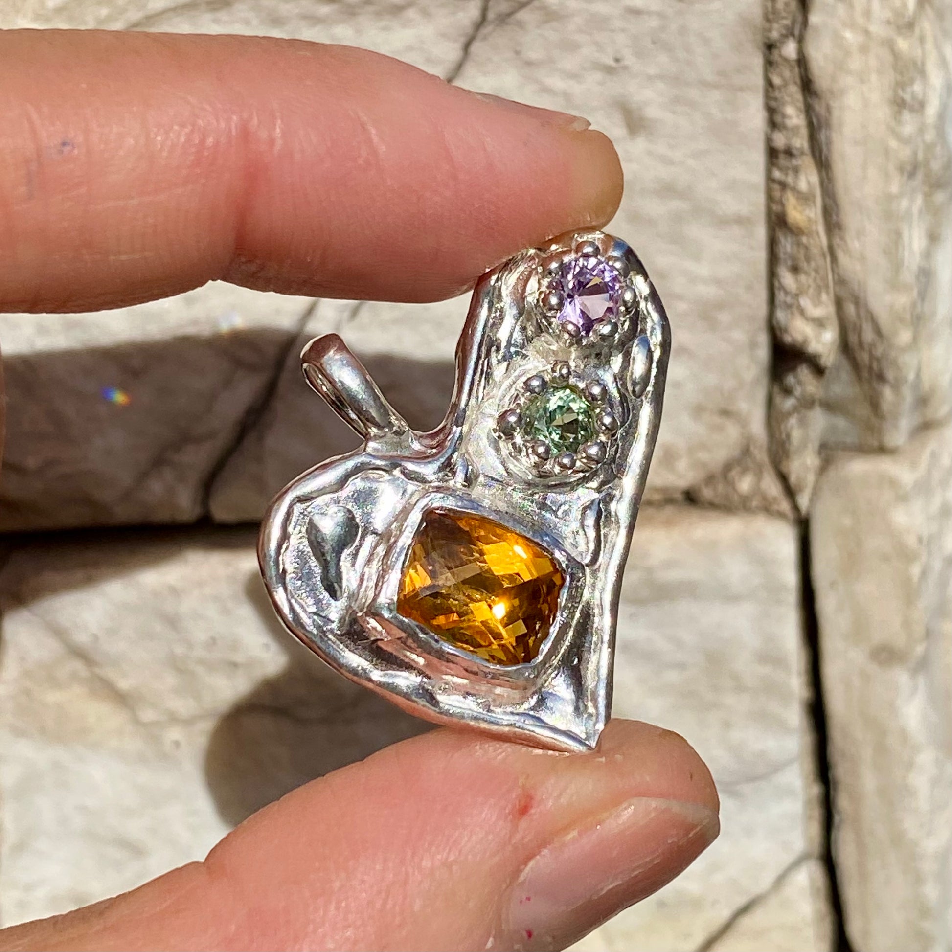 Handmade by Lia from Lia Makes Jewellery, this sterling silver heart-shaped pendant features a unique combination of colourful stones like tourmalines, sapphires, citrine, pink and blue topaz, and tiny synthetic gems, shown in a close-up to highlight the vibrant setting.
