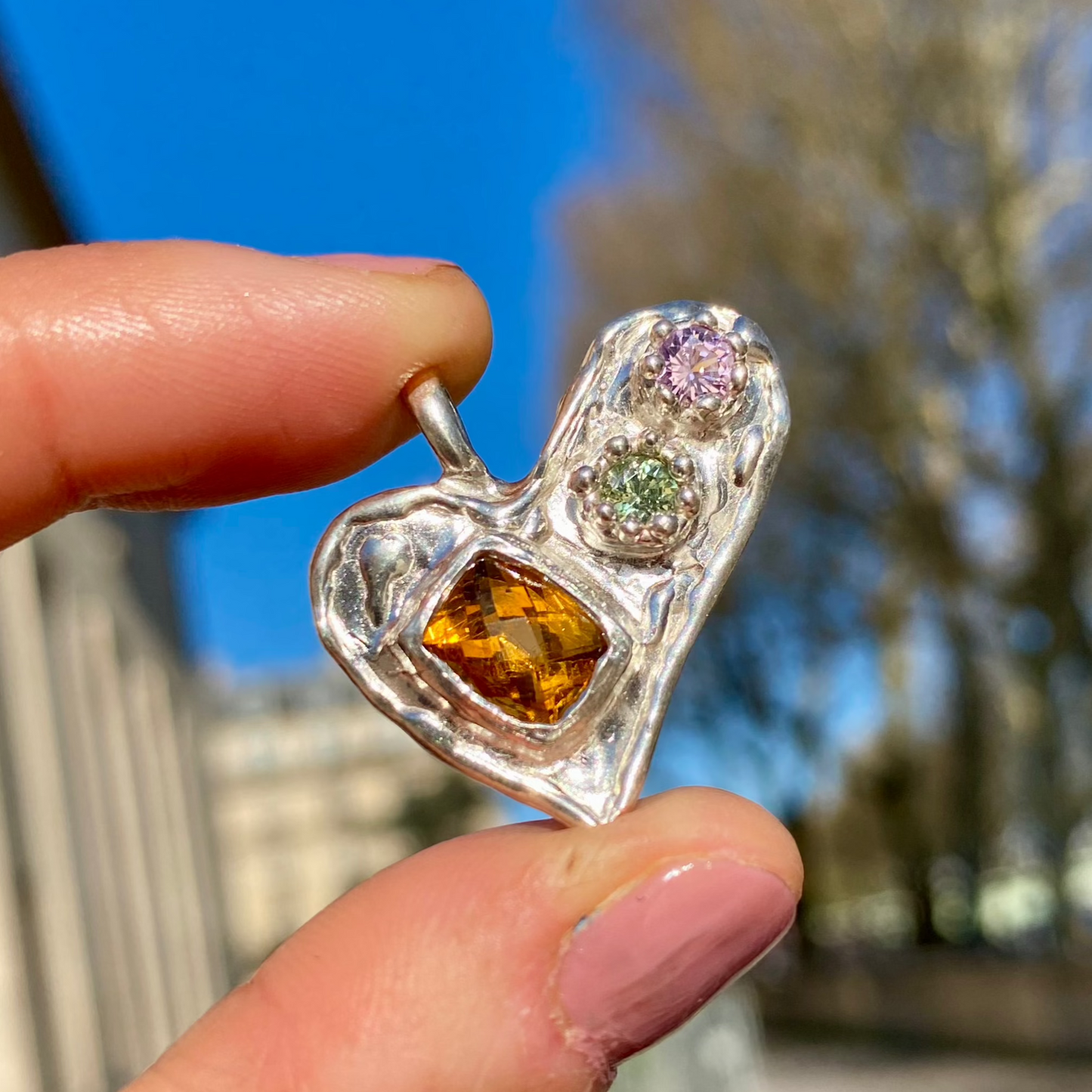 Handmade by Lia from Lia Makes Jewellery, this sterling silver heart-shaped pendant features a unique combination of colourful stones like tourmalines, sapphires, citrine, pink and blue topaz, and tiny synthetic gems, shown in a close-up to highlight the vibrant setting.
