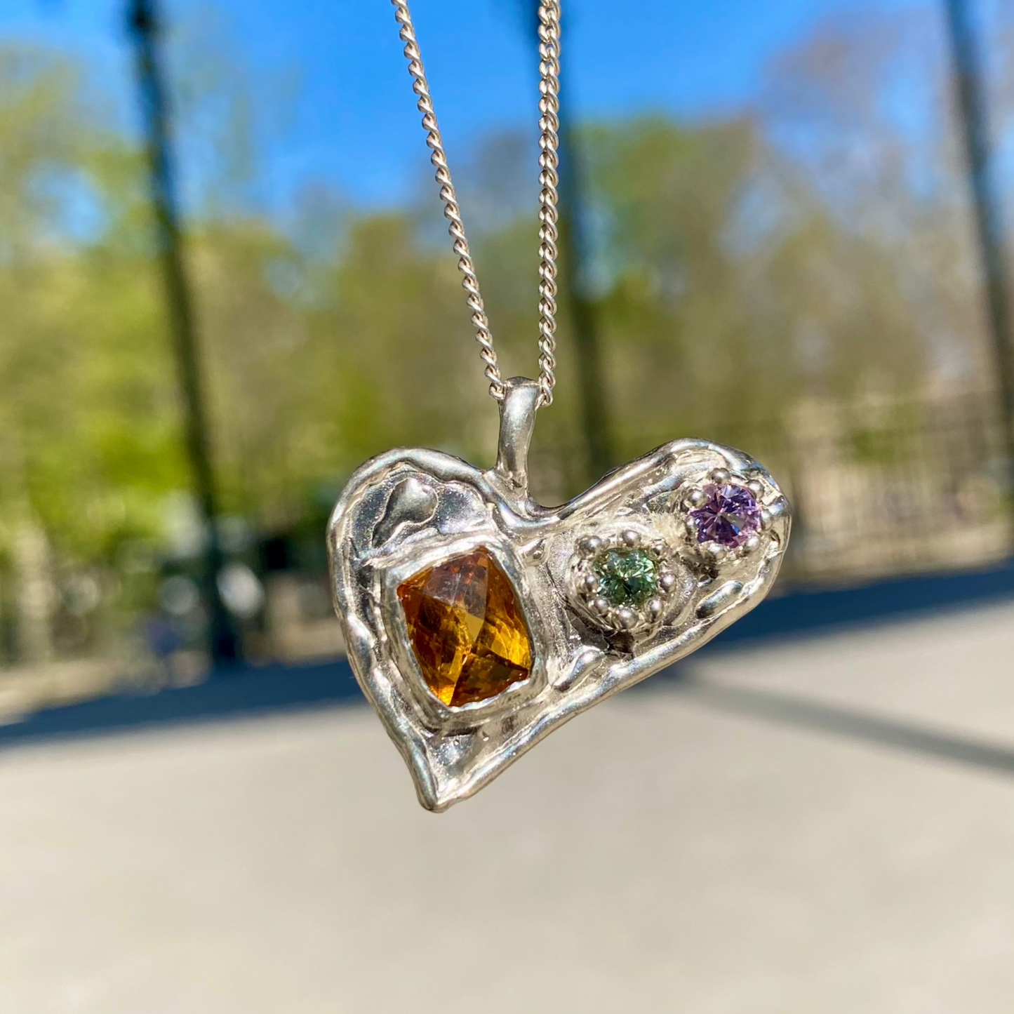 Handmade by Lia from Lia Makes Jewellery, this sterling silver heart-shaped pendant features a unique combination of colourful stones like tourmalines, sapphires, citrine, pink and blue topaz, and tiny synthetic gems, shown in a close-up to highlight the vibrant setting.
