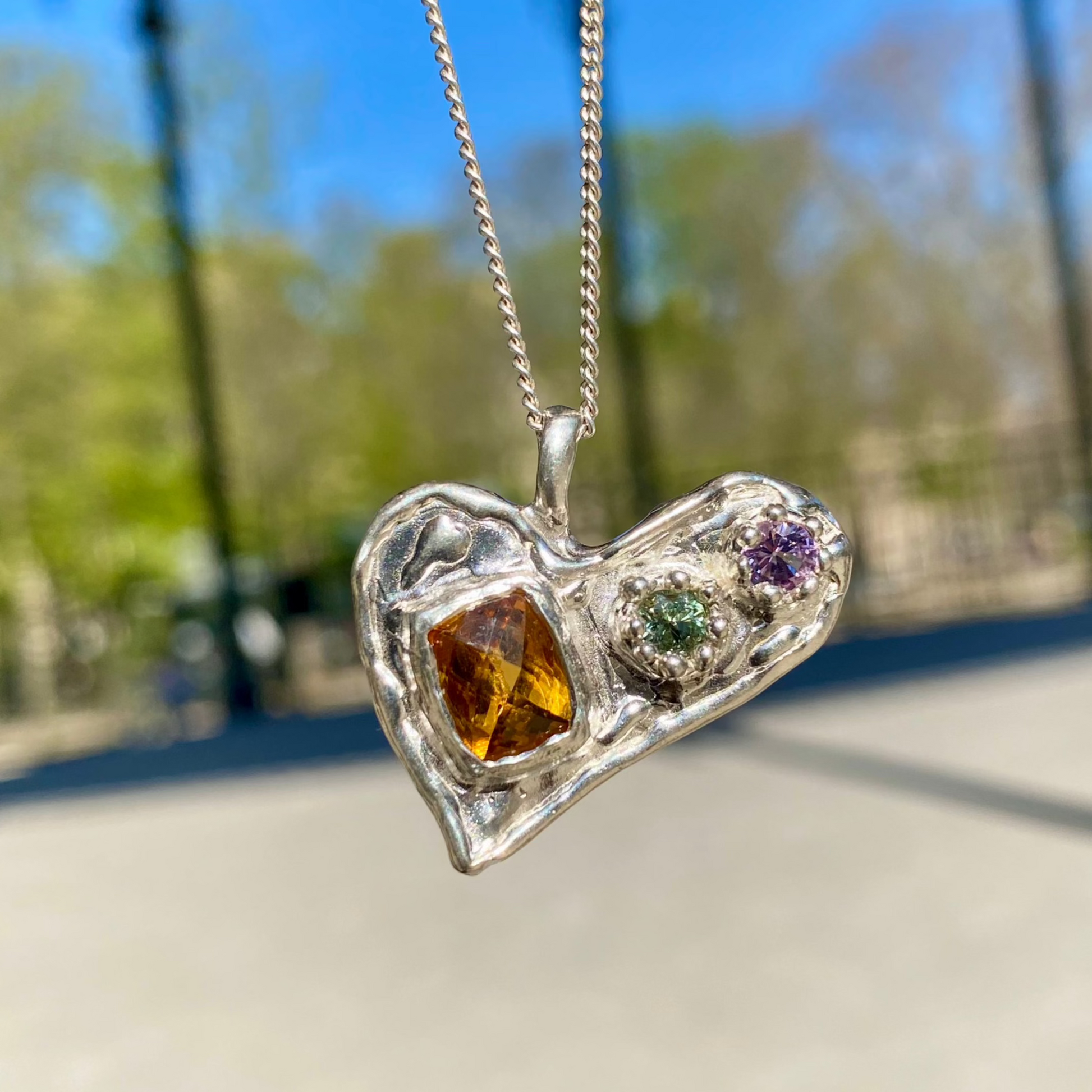 Handmade by Lia from Lia Makes Jewellery, this sterling silver heart-shaped pendant features a unique combination of colourful stones like tourmalines, sapphires, citrine, pink and blue topaz, and tiny synthetic gems, shown in a close-up to highlight the vibrant setting.

