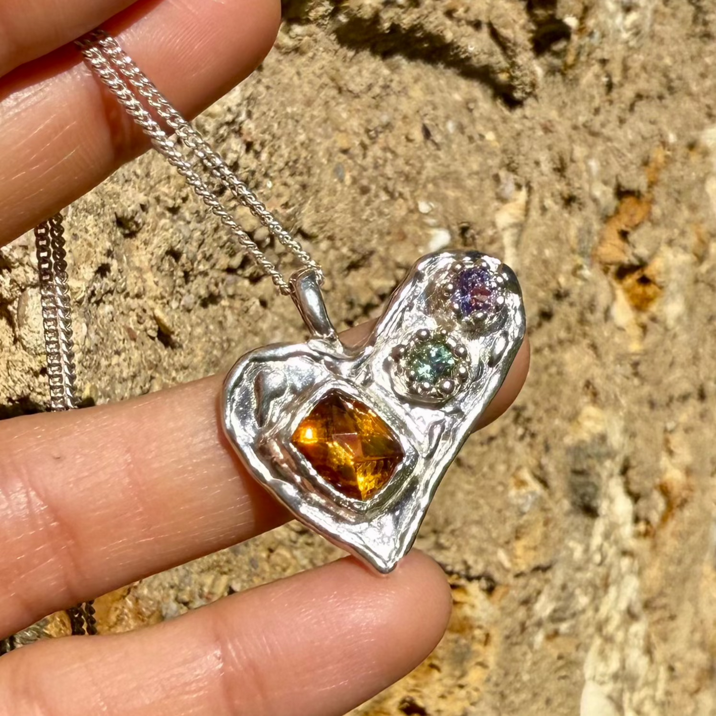 Handmade by Lia from Lia Makes Jewellery, this sterling silver heart-shaped pendant features a unique combination of colourful stones like tourmalines, sapphires, citrine, pink and blue topaz, and tiny synthetic gems, shown in a close-up to highlight the vibrant setting.
