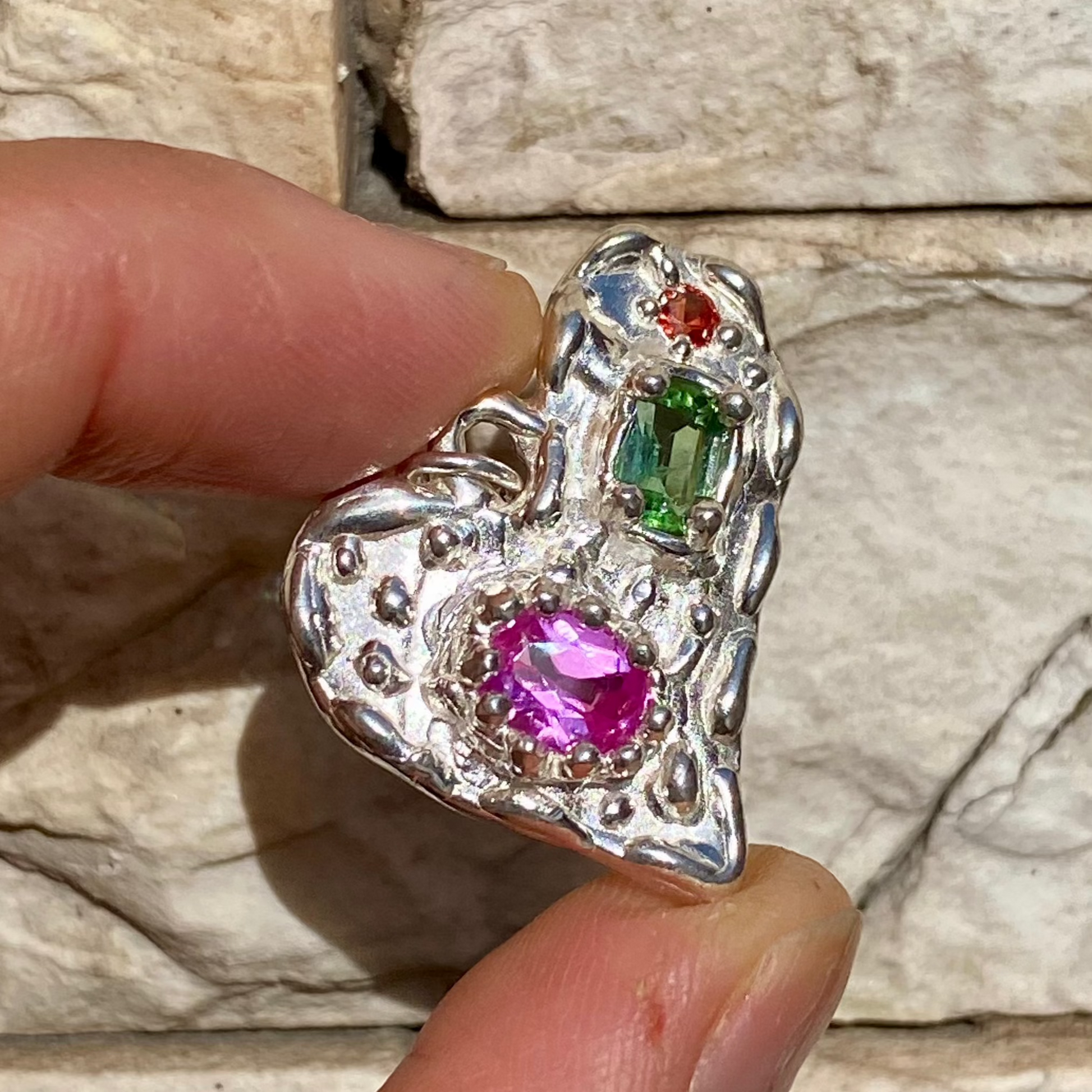 Handmade by Lia from Lia Makes Jewellery, this sterling silver heart-shaped pendant features a unique combination of colourful stones like tourmalines, sapphires, citrine, pink and blue topaz, and tiny synthetic gems, shown in a close-up to highlight the vibrant setting.
