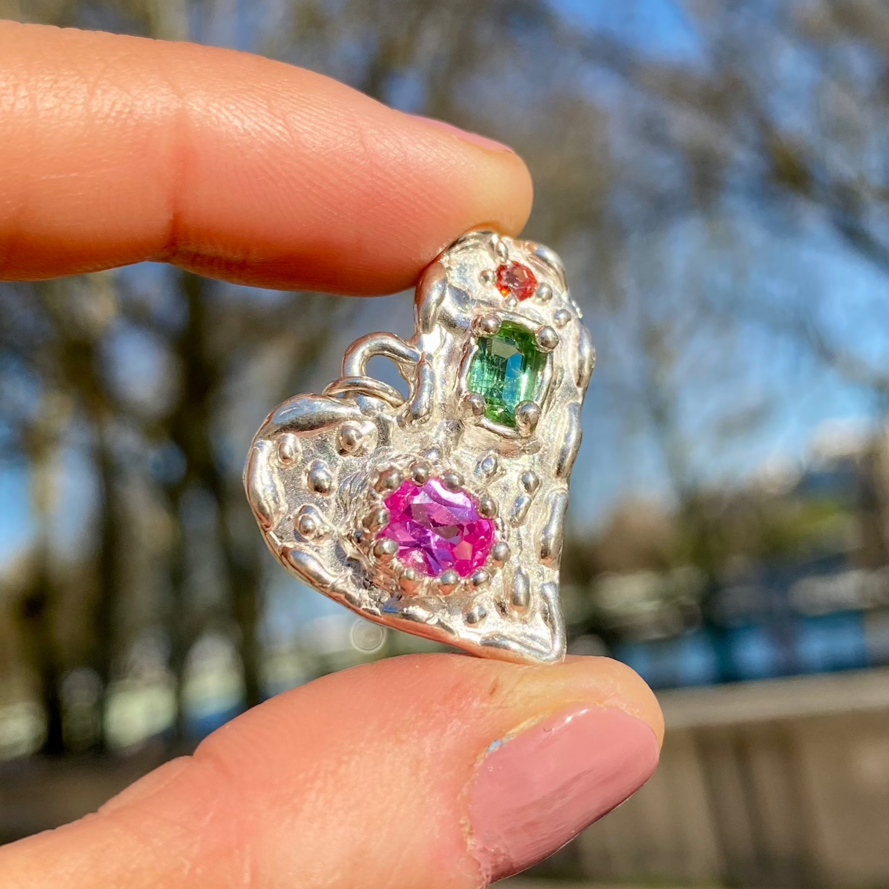 Handmade by Lia from Lia Makes Jewellery, this sterling silver heart-shaped pendant features a unique combination of colourful stones like tourmalines, sapphires, citrine, pink and blue topaz, and tiny synthetic gems, shown in a close-up to highlight the vibrant setting.
