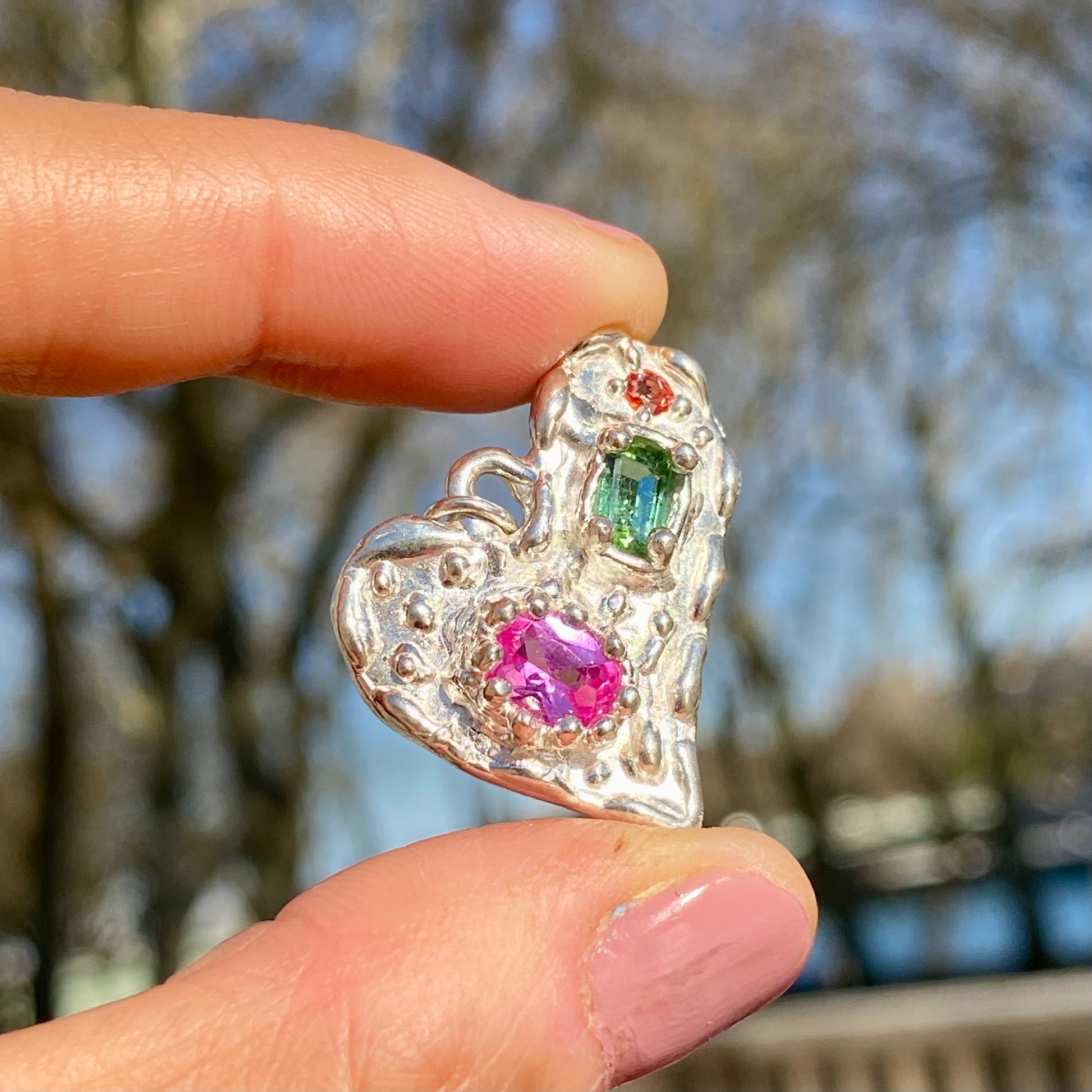 Handmade by Lia from Lia Makes Jewellery, this sterling silver heart-shaped pendant features a unique combination of colourful stones like tourmalines, sapphires, citrine, pink and blue topaz, and tiny synthetic gems, shown in a close-up to highlight the vibrant setting.
