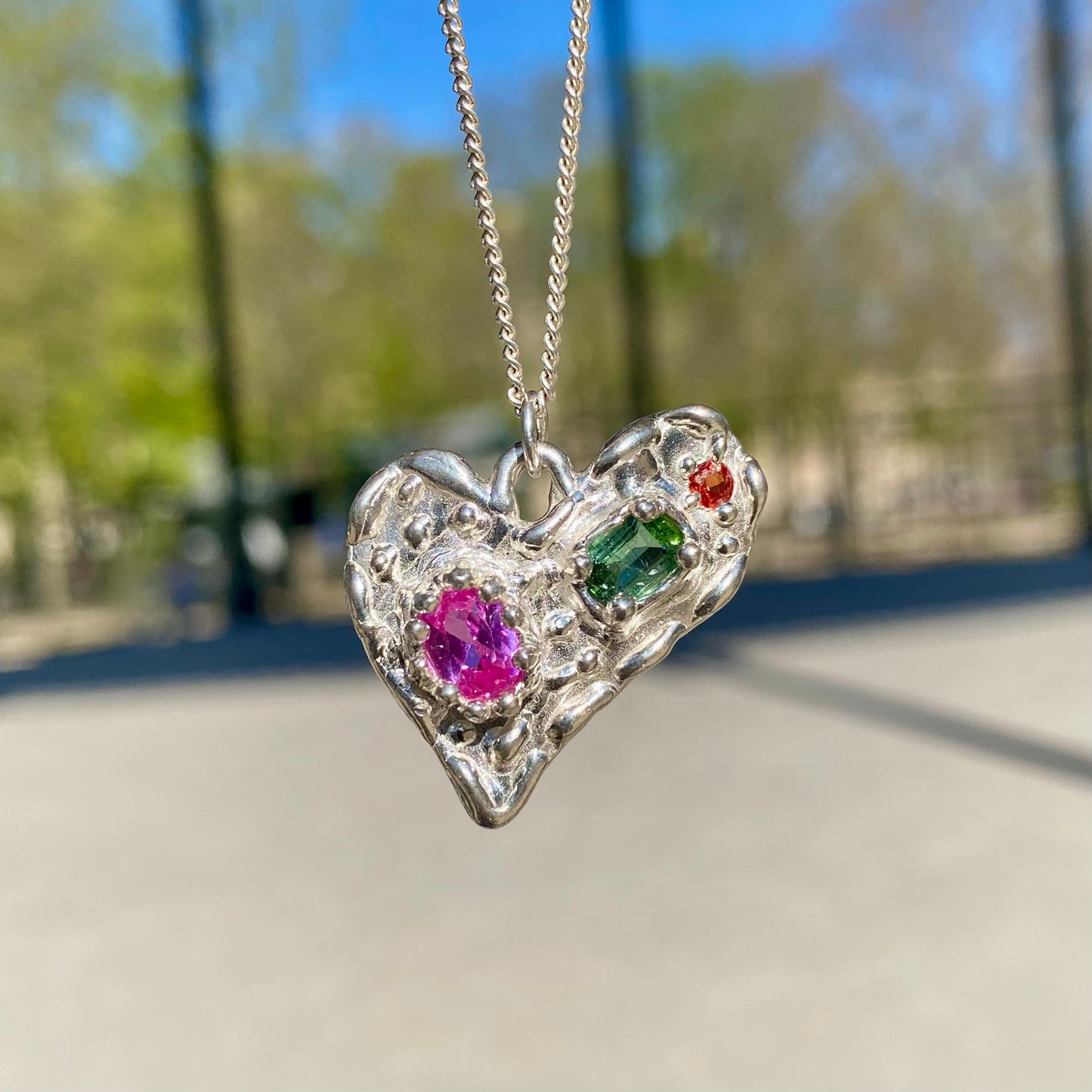 Handmade by Lia from Lia Makes Jewellery, this sterling silver heart-shaped pendant features a unique combination of colourful stones like tourmalines, sapphires, citrine, pink and blue topaz, and tiny synthetic gems, shown in a close-up to highlight the vibrant setting.
