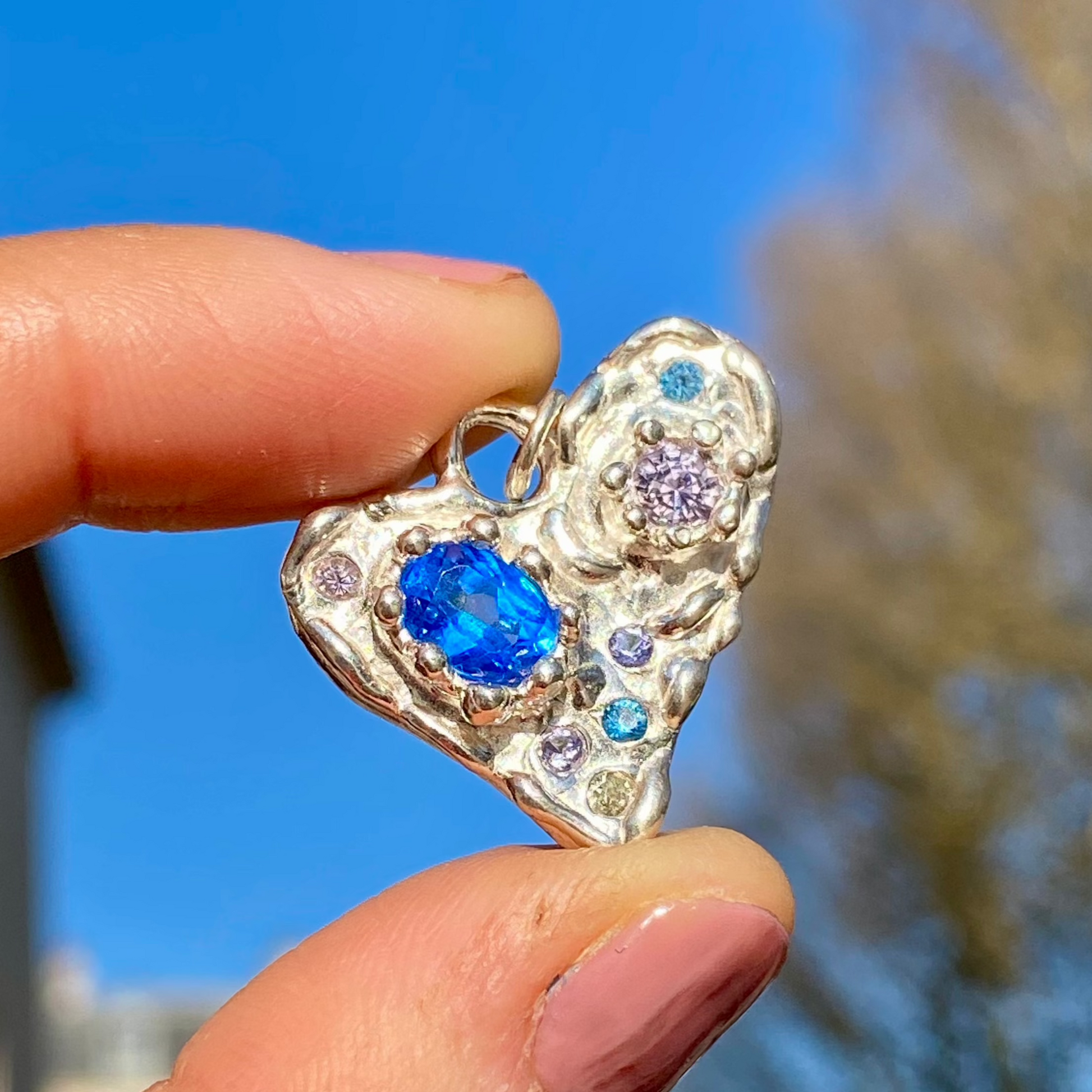Handmade by Lia from Lia Makes Jewellery, this sterling silver heart-shaped pendant features a unique combination of colourful stones like tourmalines, sapphires, citrine, pink and blue topaz, and tiny synthetic gems, shown in a close-up to highlight the vibrant setting.
