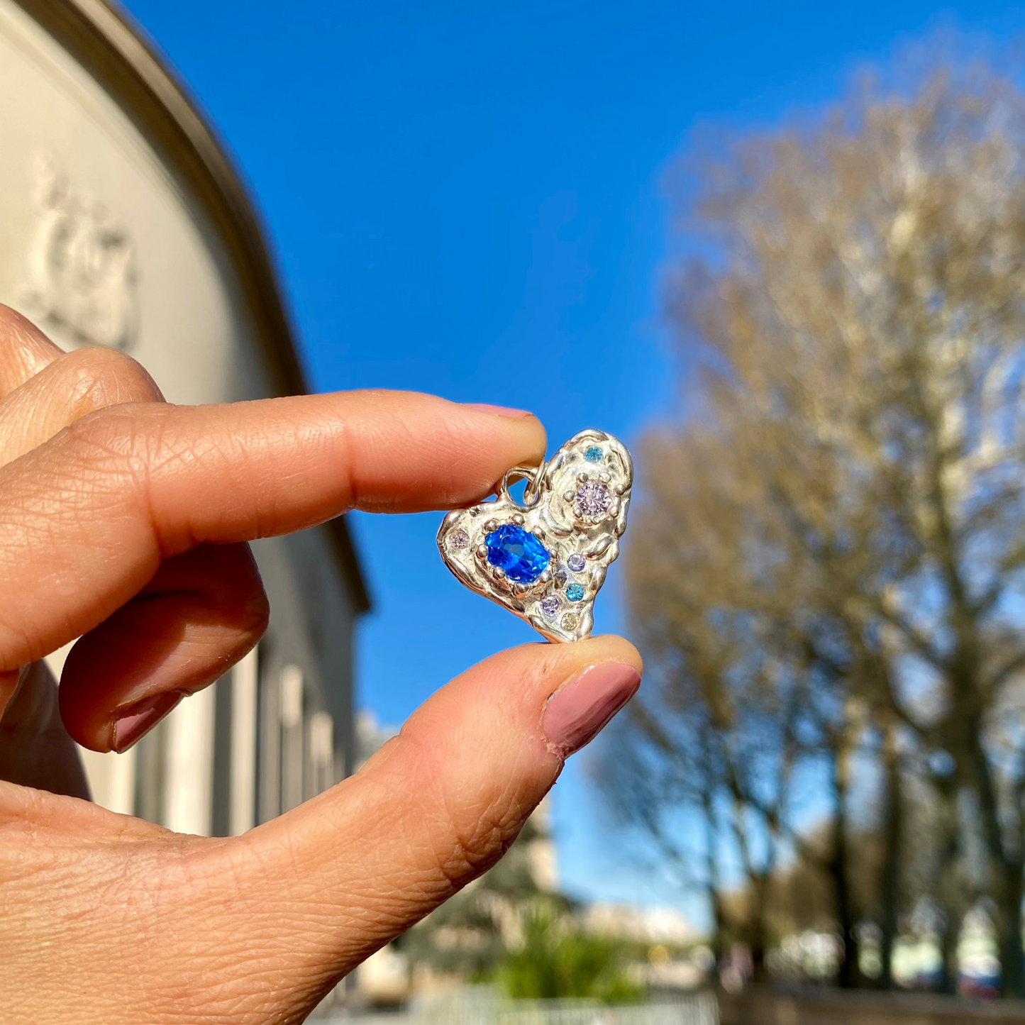 Handmade by Lia from Lia Makes Jewellery, this sterling silver heart-shaped pendant features a unique combination of colourful stones like tourmalines, sapphires, citrine, pink and blue topaz, and tiny synthetic gems, shown in a close-up to highlight the vibrant setting.
