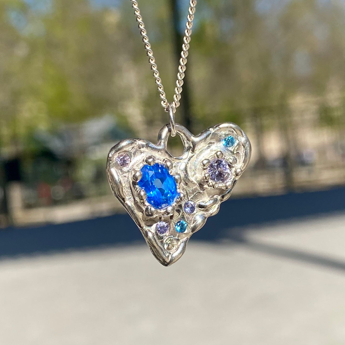 Handmade by Lia from Lia Makes Jewellery, this sterling silver heart-shaped pendant features a unique combination of colourful stones like tourmalines, sapphires, citrine, pink and blue topaz, and tiny synthetic gems, shown in a close-up to highlight the vibrant setting.
