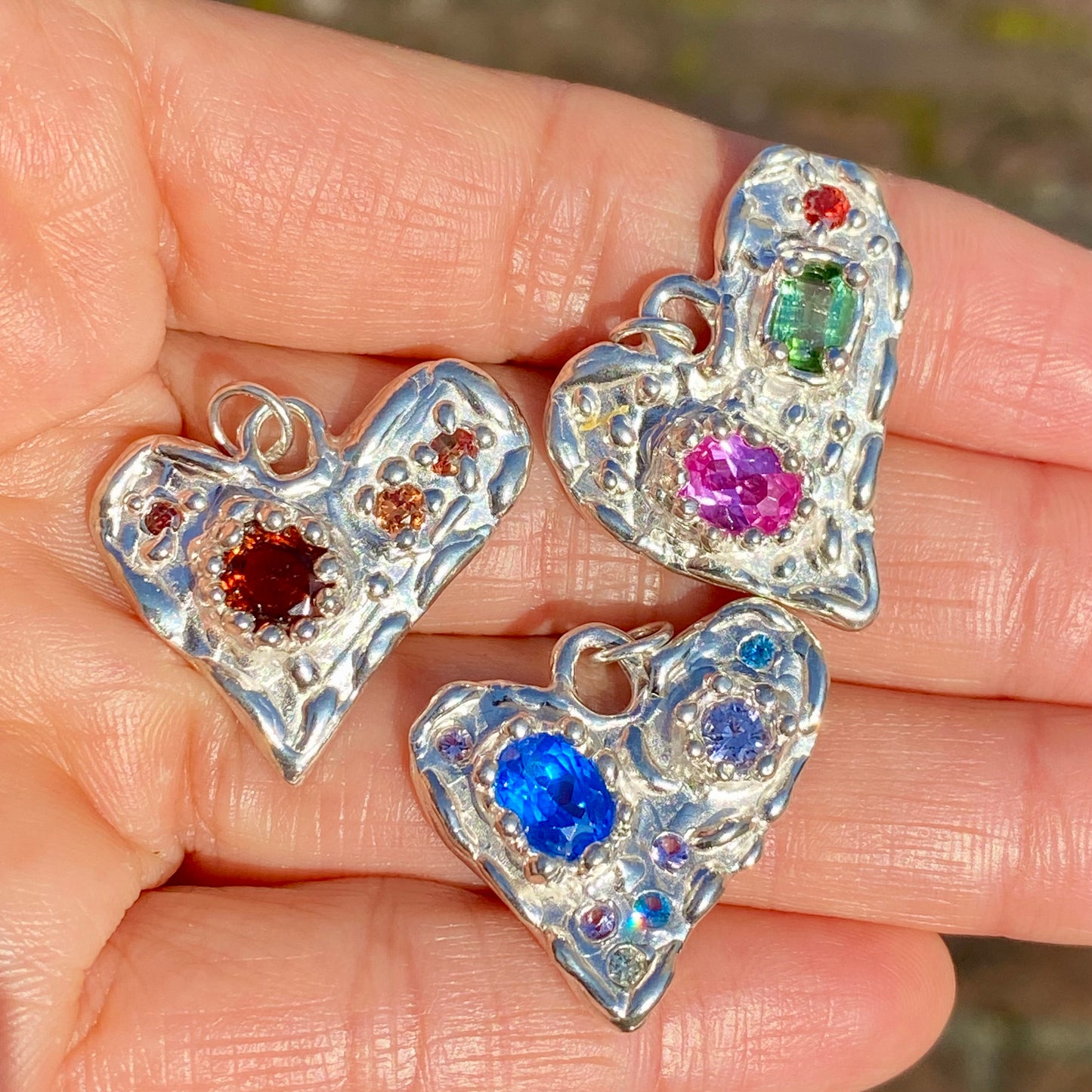 Handmade by Lia from Lia Makes Jewellery, this sterling silver heart-shaped pendant features a unique combination of colourful stones like tourmalines, sapphires, citrine, pink and blue topaz, and tiny synthetic gems, shown in a close-up to highlight the vibrant setting.
