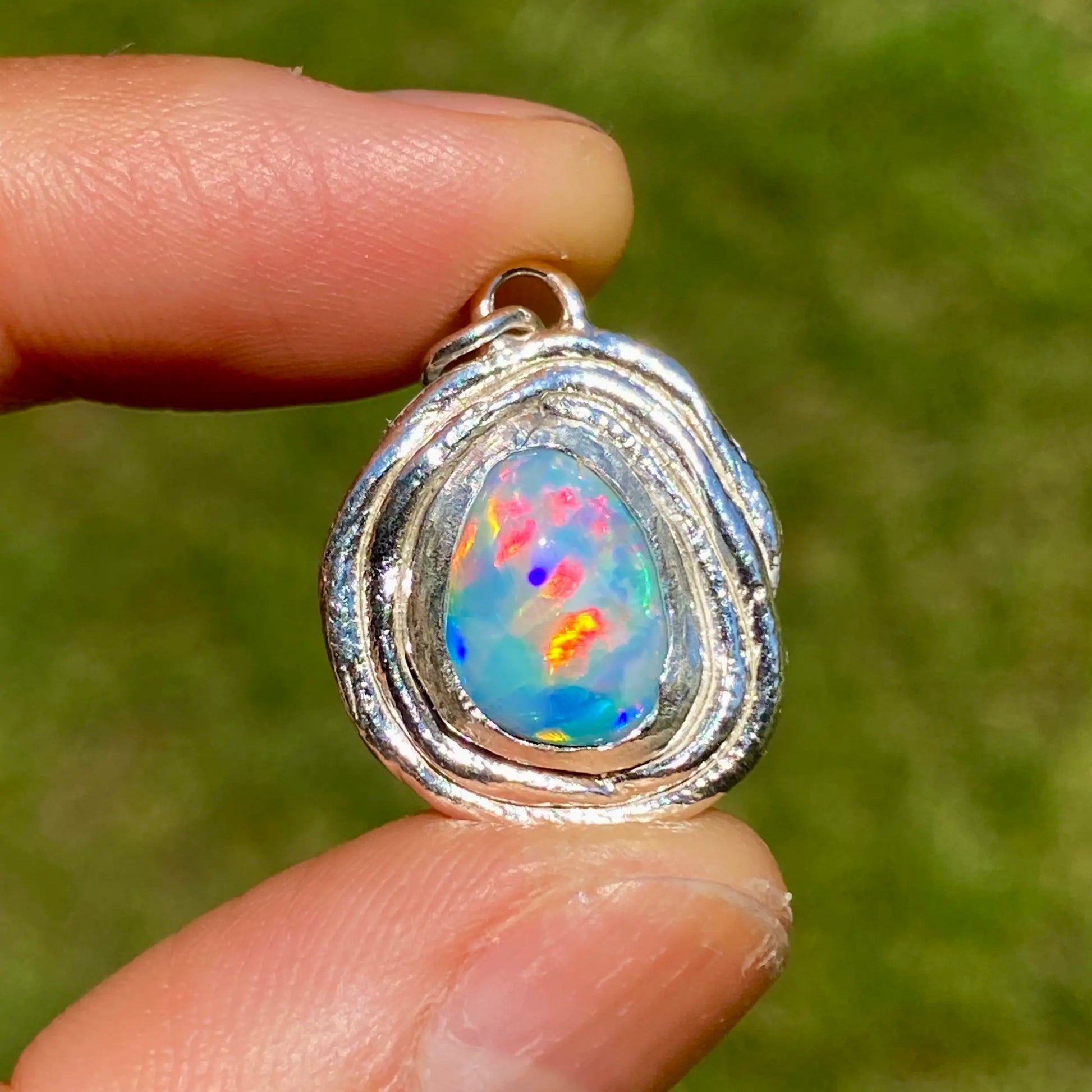 Opal pendant in a silver setting held between two fingers against a blurred green background