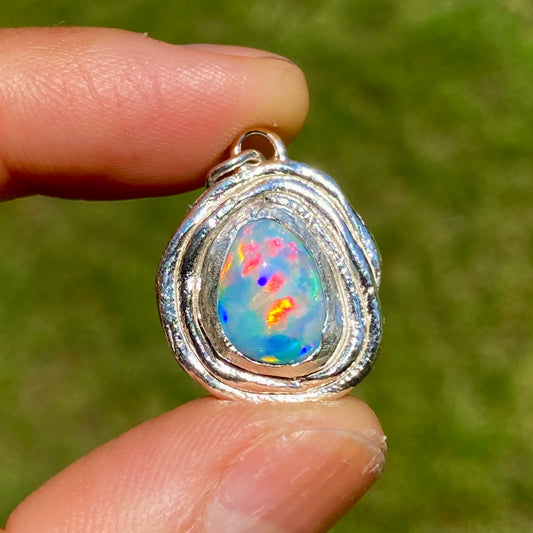 Opal pendant in a silver setting held between two fingers against a blurred green background