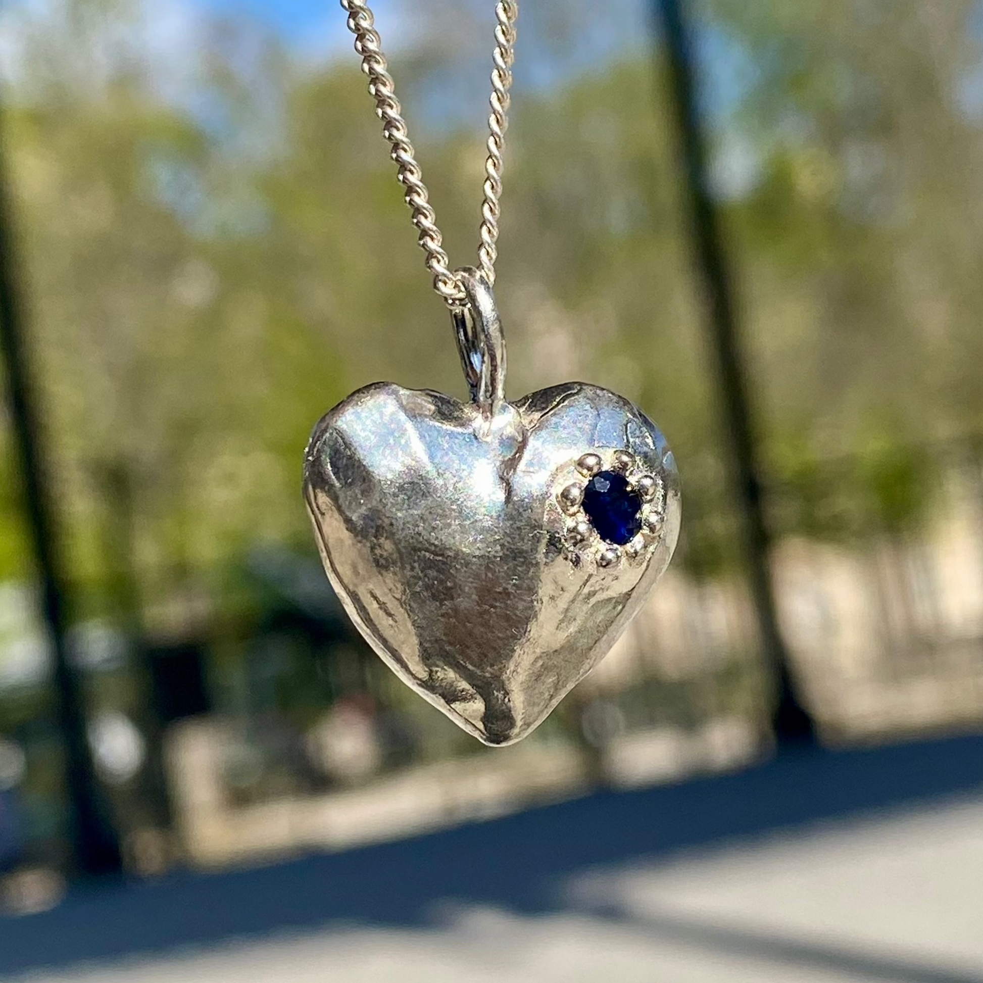 The Petite Heart pendant is a heart-shaped piece, handmade by me in sterling silver. Each pendant features a sapphire stone and is available in four colours: yellow, pink, orange, or blue.