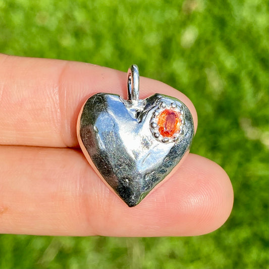 The Petite Heart pendant is a heart-shaped piece, handmade by me in sterling silver. Each pendant features a sapphire stone and is available in four colours: yellow, pink, orange, or blue.