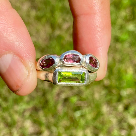 The Spring Blossom ring stack comes as a set of two. One ring features three pink tourmaline stones, and the other has a long green rectangular peridot stone. Handmade from sterling silver, these stacking rings are available in one size: O ½ (US 7 ¼).