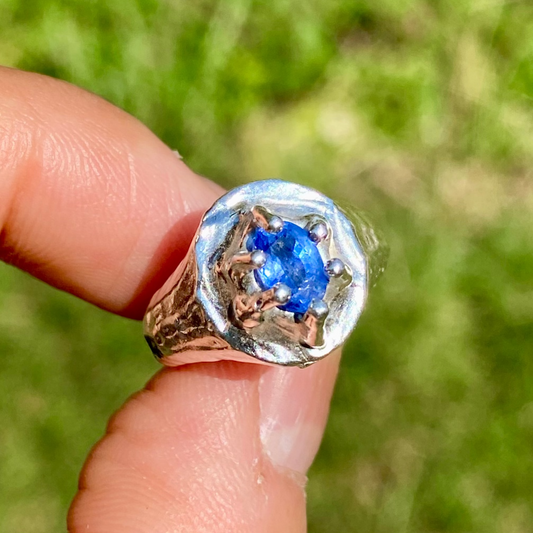 Silver ring with a blue gemstone held on a finger against a blurred green background