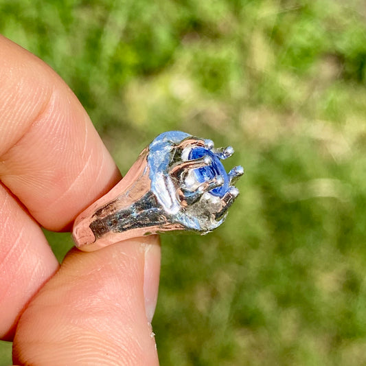 Silver ring with a blue stone held between two fingers against a blurred green background
